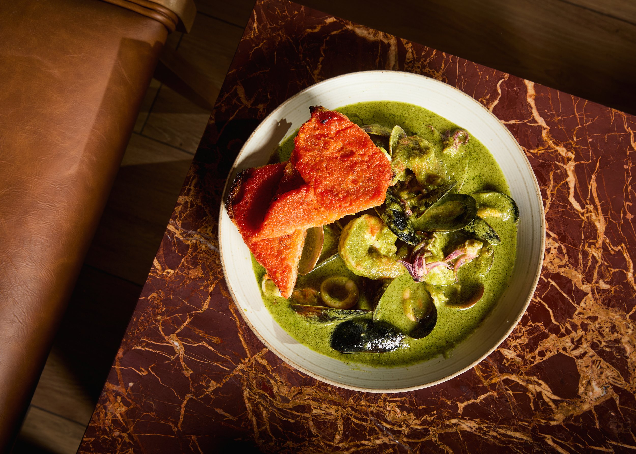 A bowl of green seafood curry with mussels and vegetables, served with fried breaded fish on top, on a marble table.