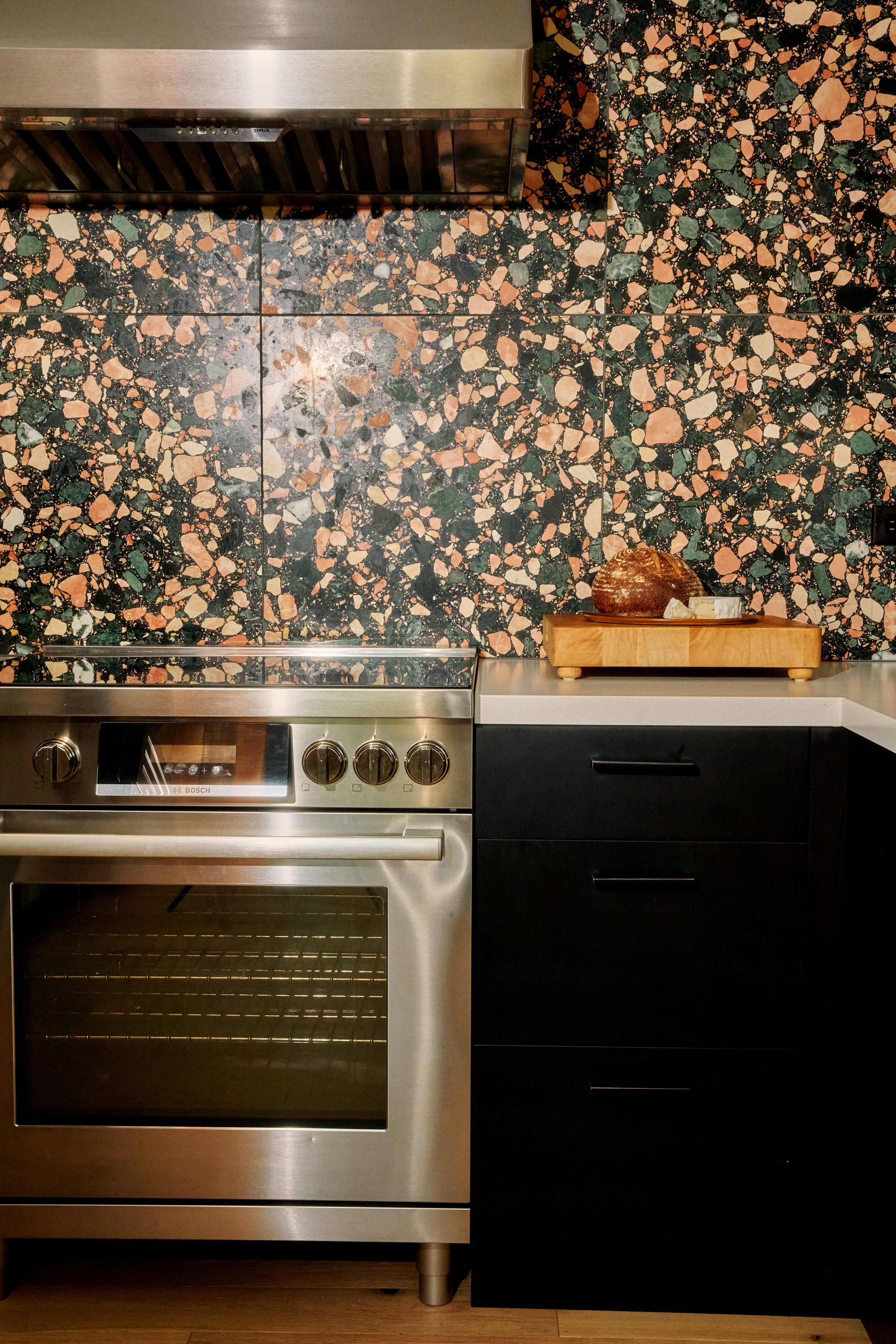 Stainless steel oven with control knobs in a kitchen with multicolored terrazzo backsplash and dark cabinet to the right, featuring a wooden tray with bread and butter on top.
