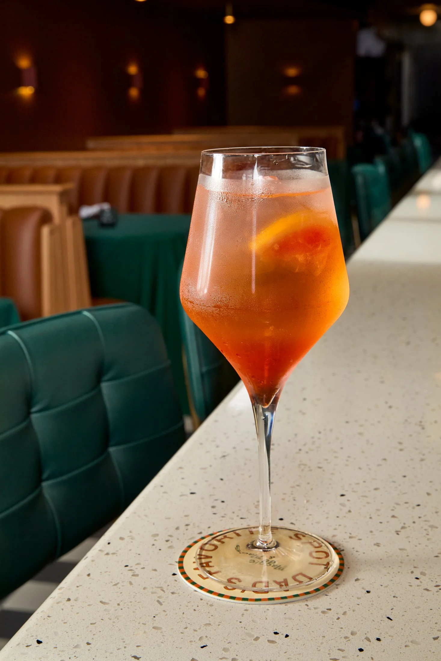A pink cocktail with an orange slice garnish in a tall, stemmed glass on a bar counter.