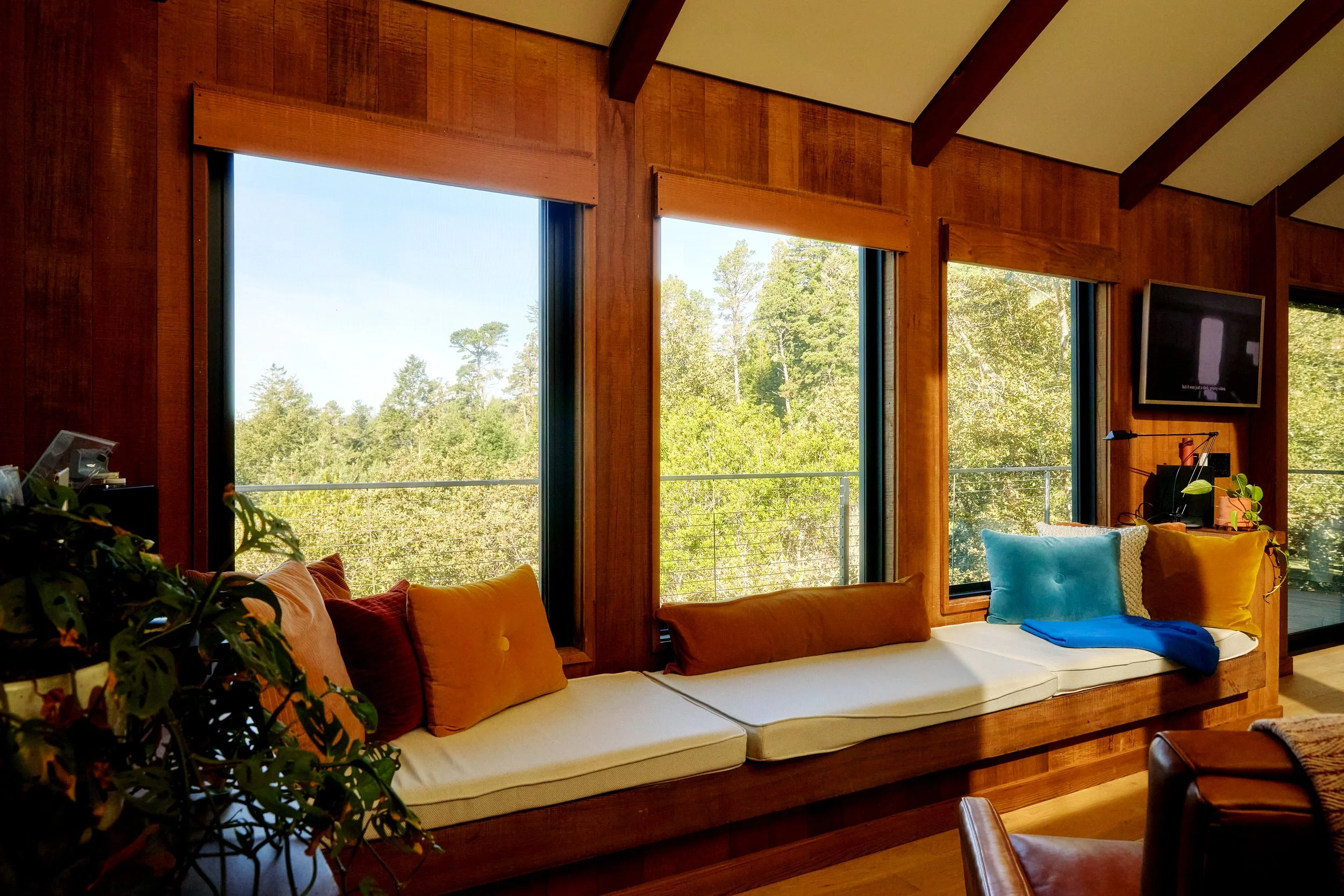 Living room with large windows showing greenery outside, built-in wooden window seat with colorful pillows, and a mounted TV on the wall.