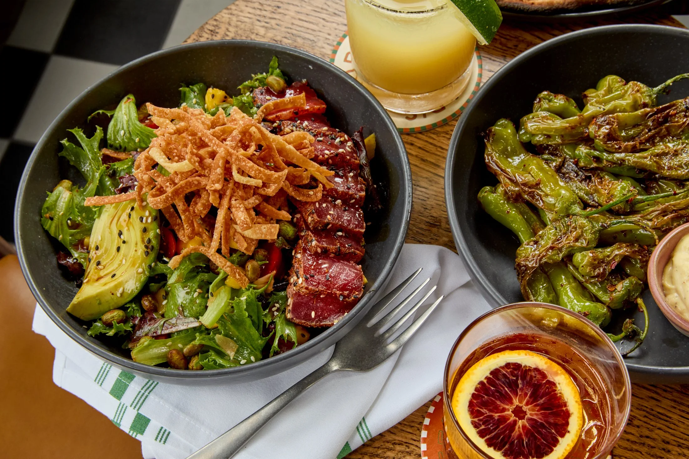 A black bowl filled with mixed greens, sliced avocado, cherry tomatoes, shredded cheese, sesame seeds, and pieces of seared tuna, accompanied by a tall glass of yellowish cocktail with lime wedge, and a side dish of grilled shishito peppers, garnishe