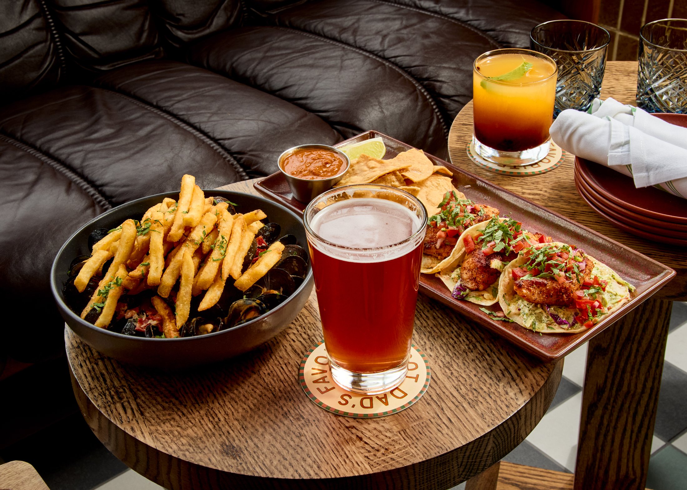 A wooden table with a serving of French fries, a bowl of salsa, and a plate of tacos topped with salsa and chopped herbs. There are two glasses of beer, one with lime, on the table. Napkins are on a plate, and a glass of water and two empty glasses a