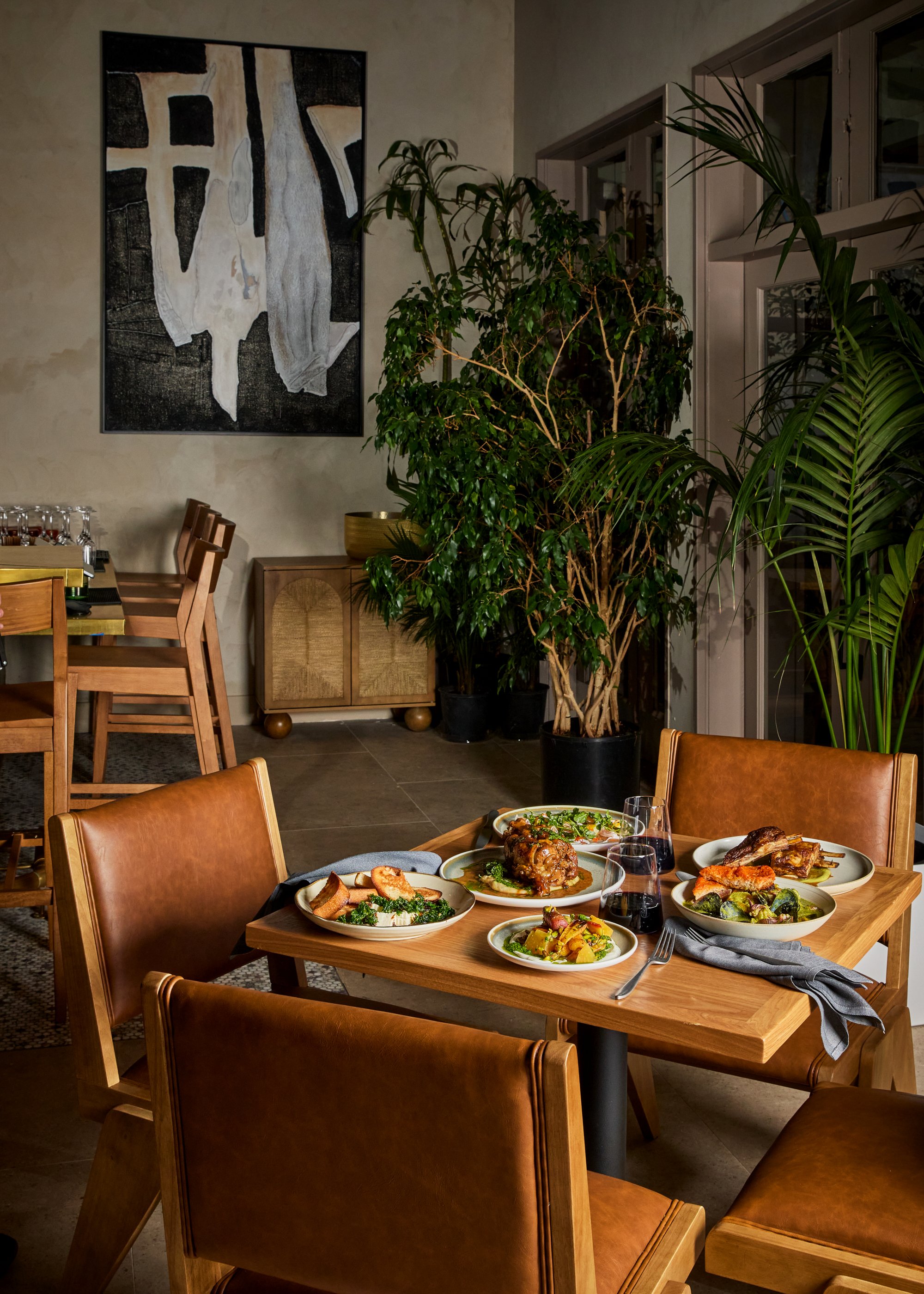 A dining table set with various plates of food, including meat and vegetable dishes, with glasses of red wine, in a cozy restaurant or home dining room, decorated with large green plants and abstract wall art.