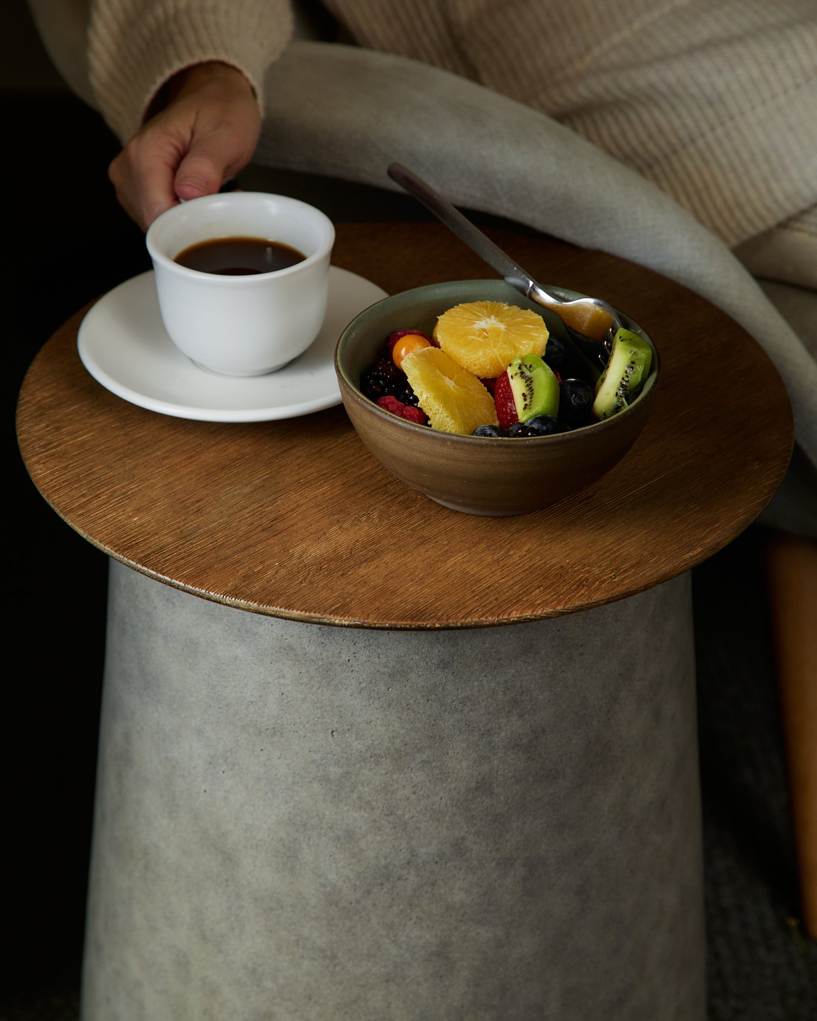 A person seated at a wooden table, holding a cup of black coffee, with a bowl of mixed fruits including orange slices, kiwi, berries, and a spoon, and wearing a beige, striped sweater.