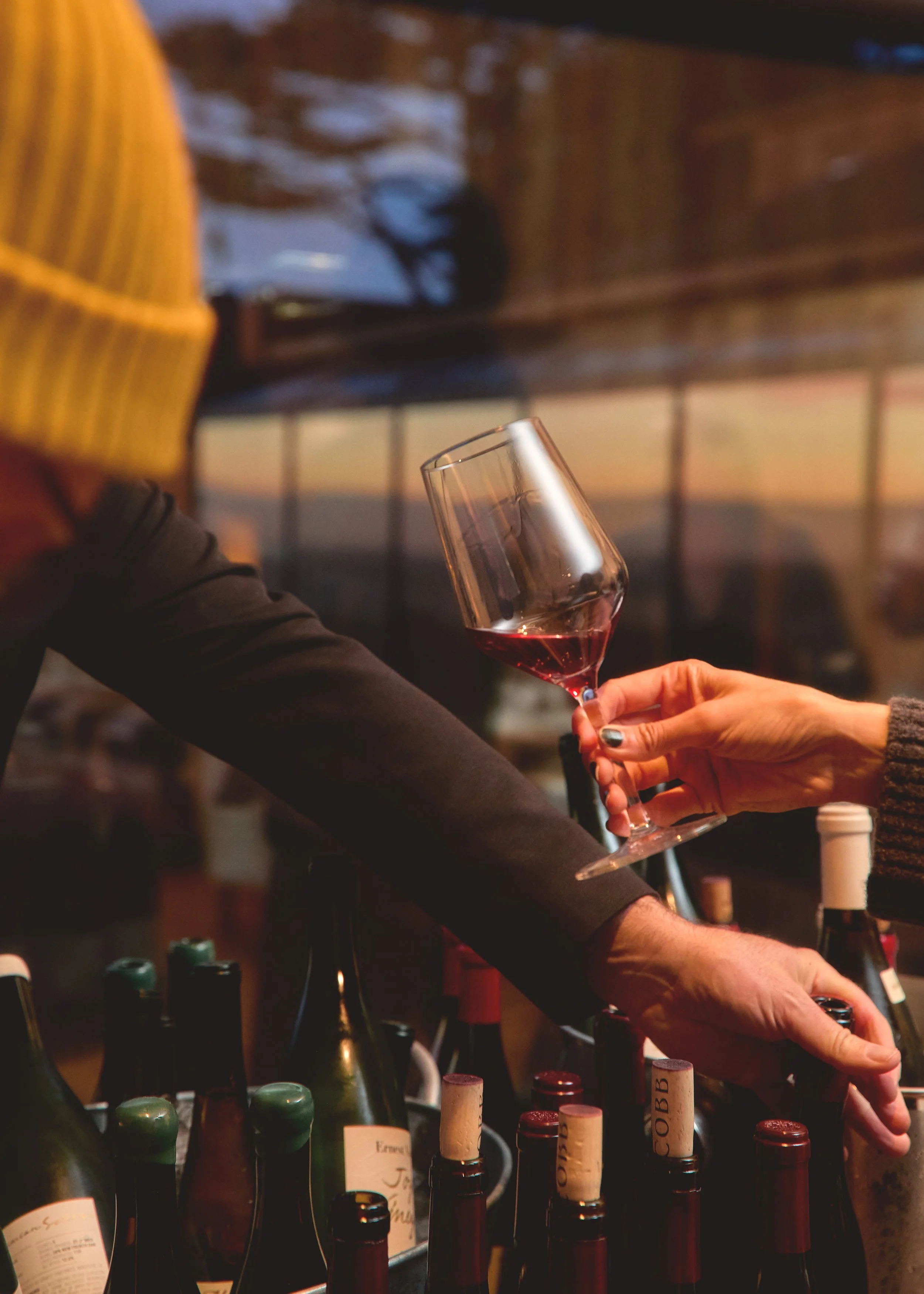 Person pouring red wine into a glass at a bar with bottles of wine in the foreground and a sunset visible through the window in the background.