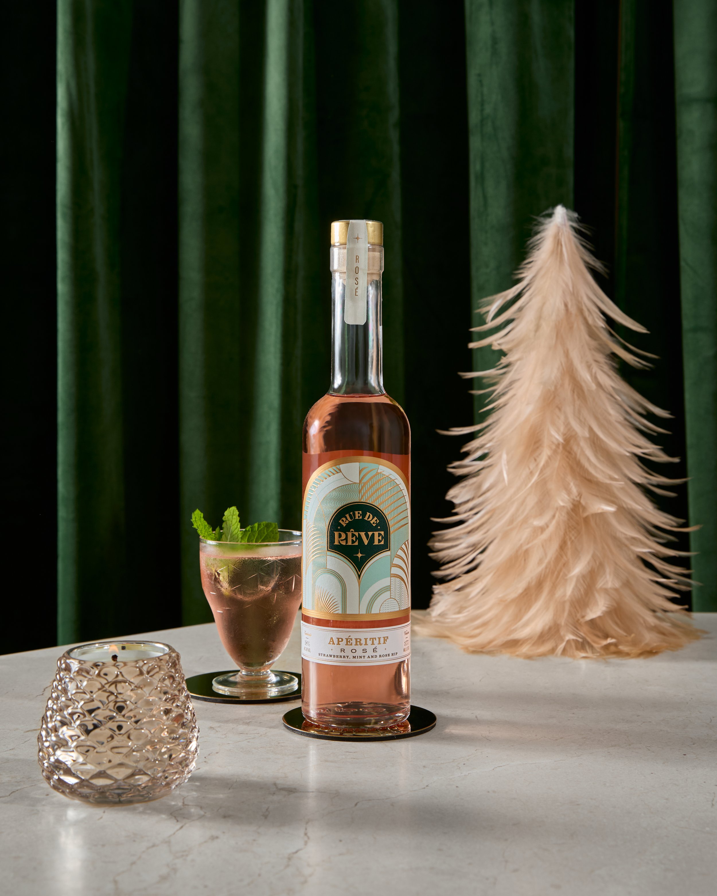 A bottle of Rue de Rêve rosé apéritif drink, a glass with pink beverage garnished with mint leaves, a decorative candle holder, and a beige feathered Christmas tree on a white tabletop with green curtains in the background.