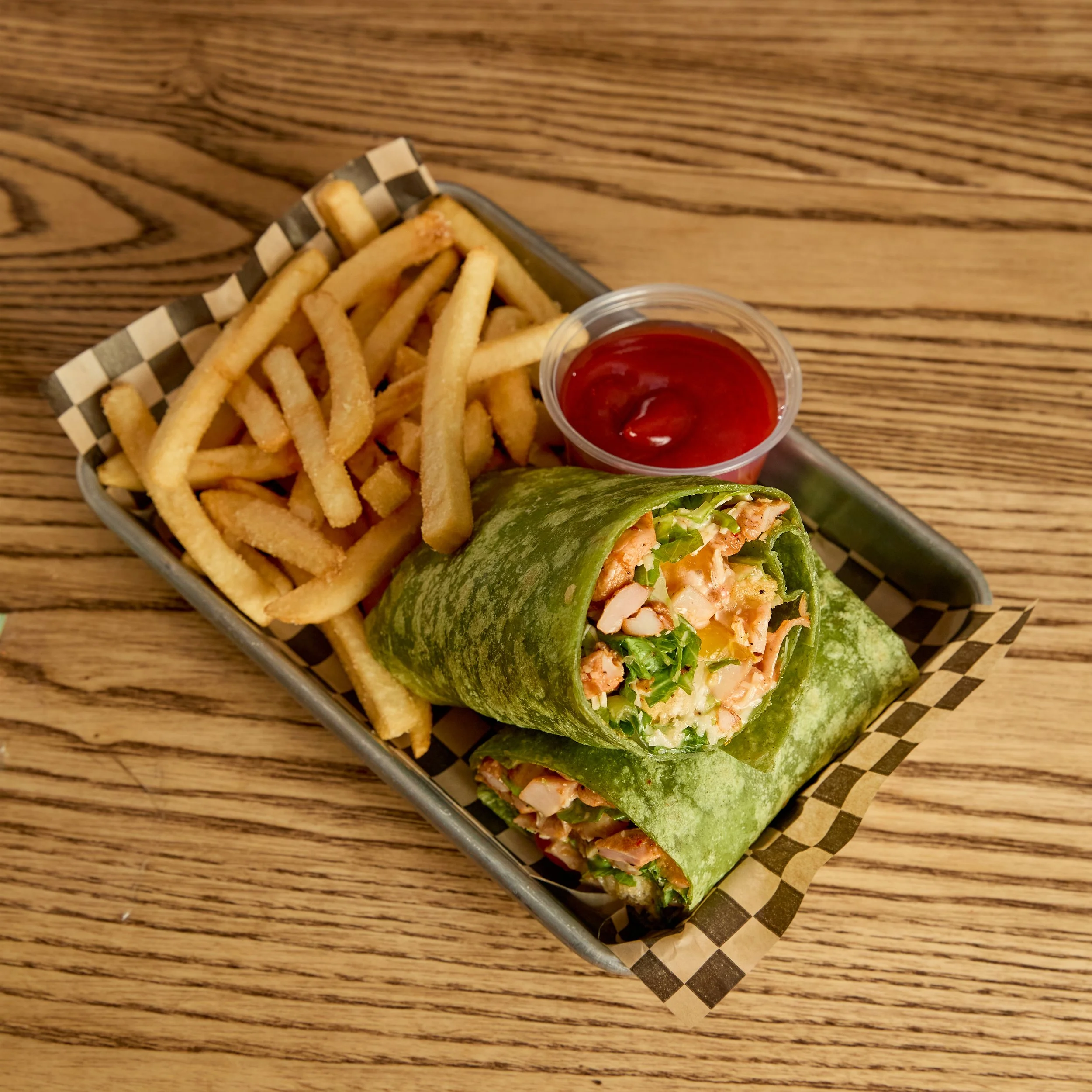 A basket with French fries, a green wrap sandwich filled with lettuce, chicken, and other ingredients, and a side of ketchup on a wooden table.