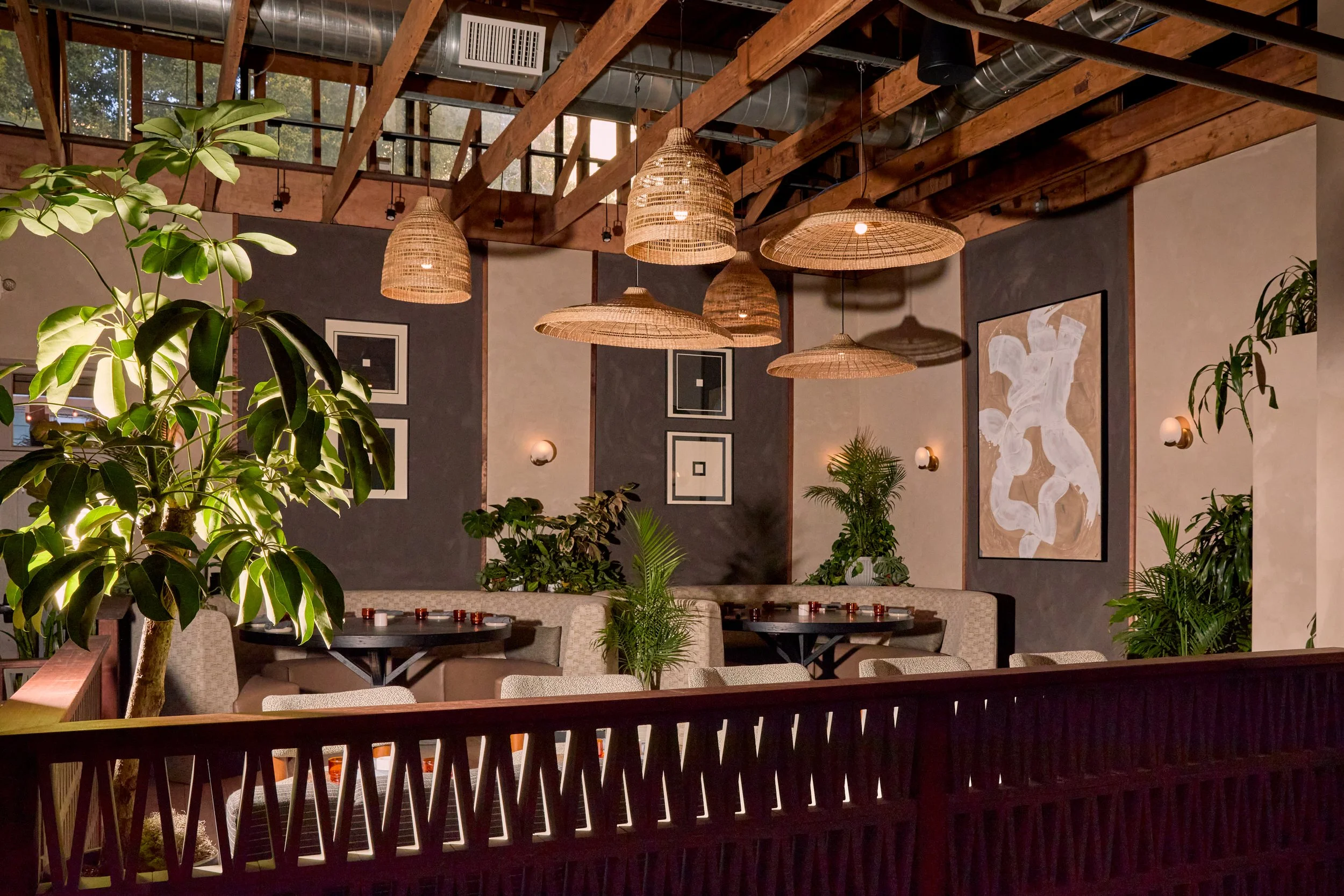Interior of a restaurant with wooden beams, hanging wicker pendant lights, framed artwork, and green plants, with tables and chairs arranged for dining.