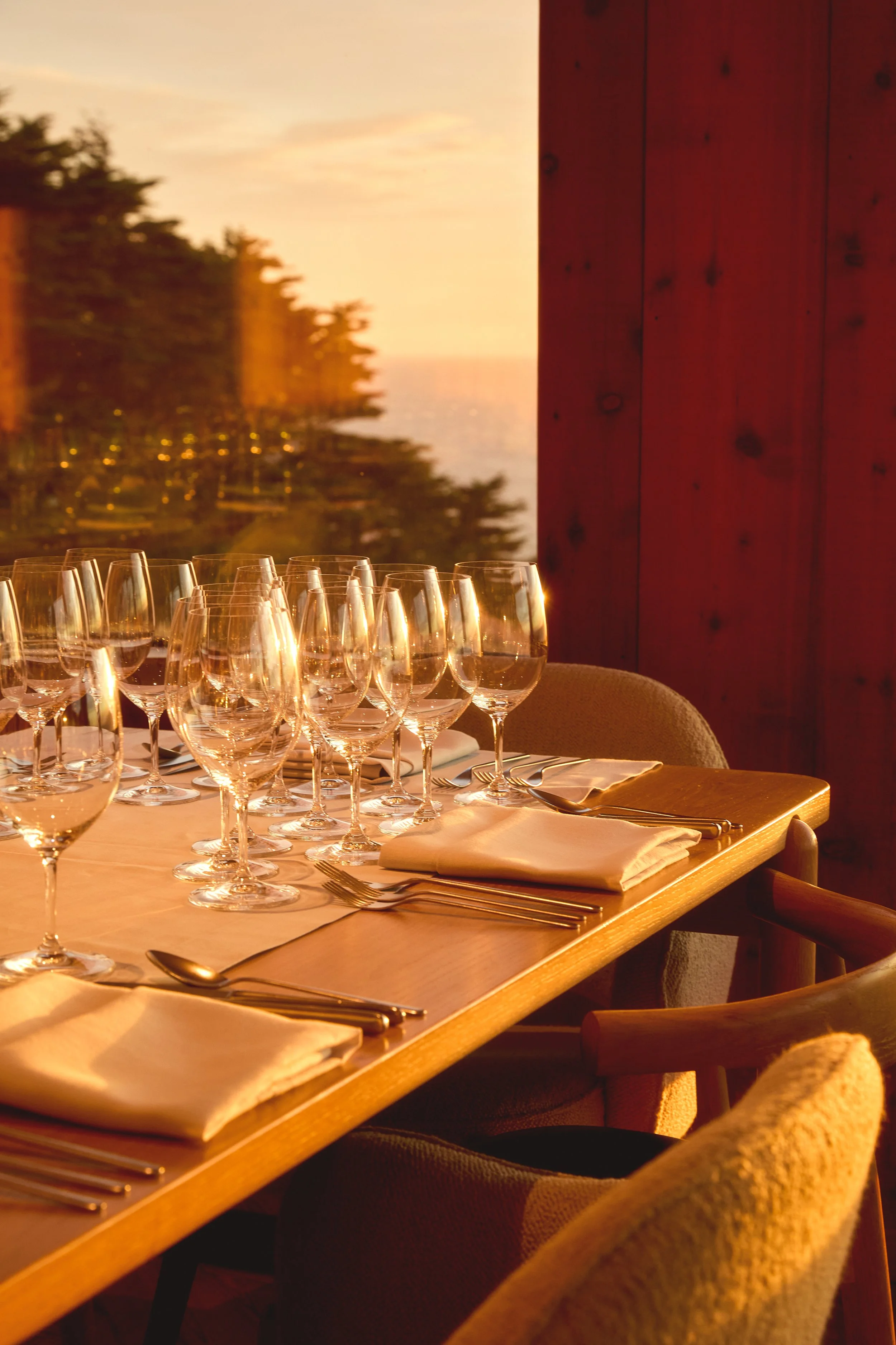A dining table set with wine glasses, napkins, and cutlery, with a sunset view of trees and the ocean through a window.