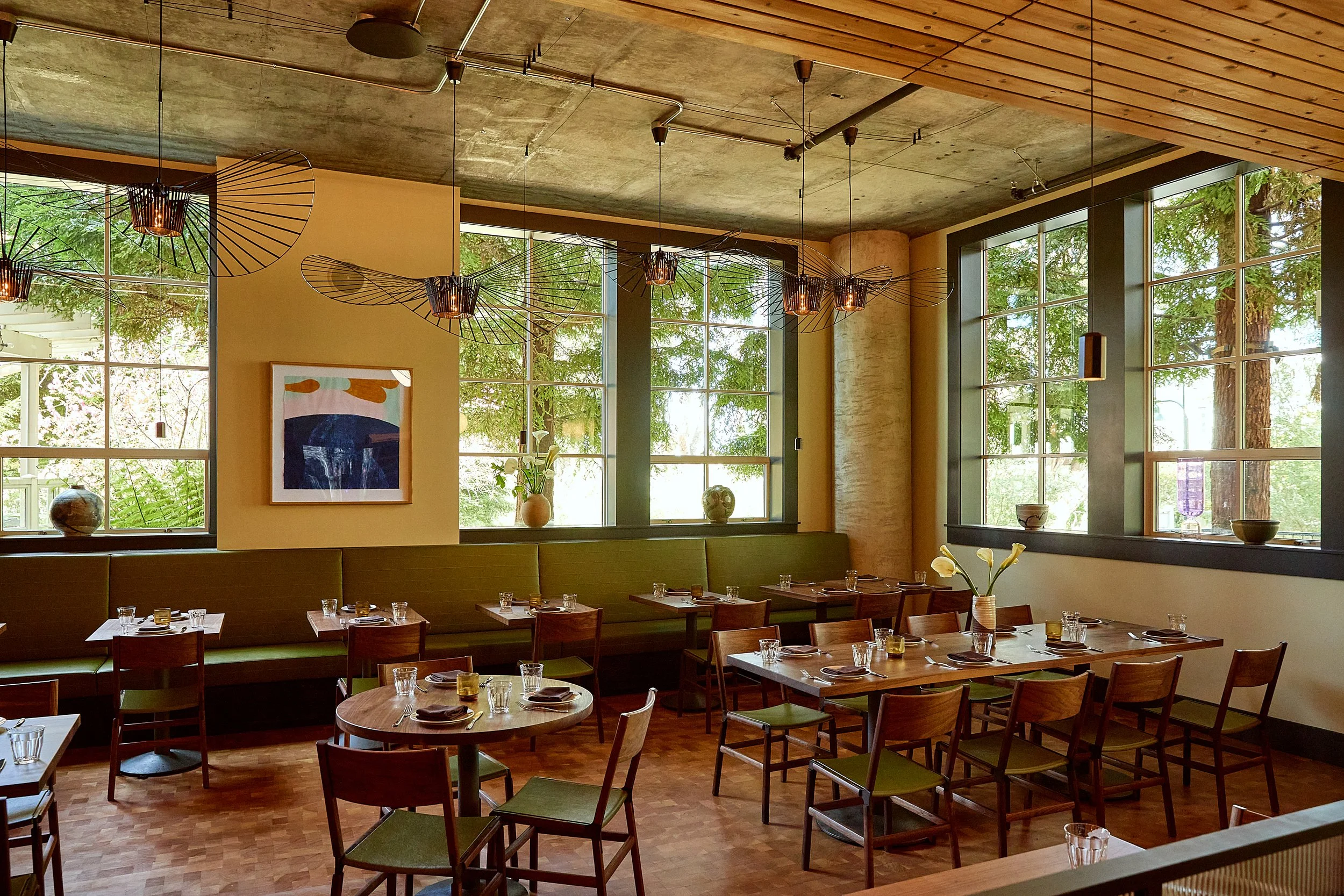 Interior of a modern restaurant with large windows, wooden tables and chairs, green cushioned seating along the wall, and decorative lighting fixtures hanging from a concrete ceiling.