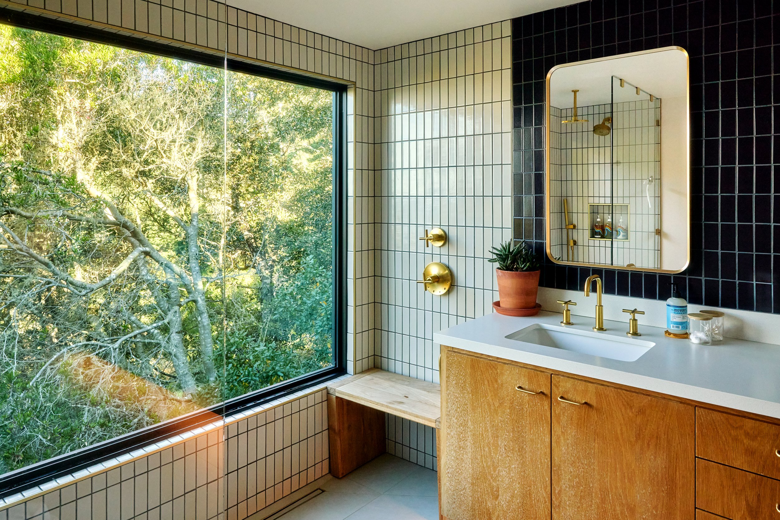 Modern bathroom with large window overlooking trees, black and white tile walls, gold fixtures, wooden vanity, white countertop, mirror, potted plant, soap, and skincare bottles.