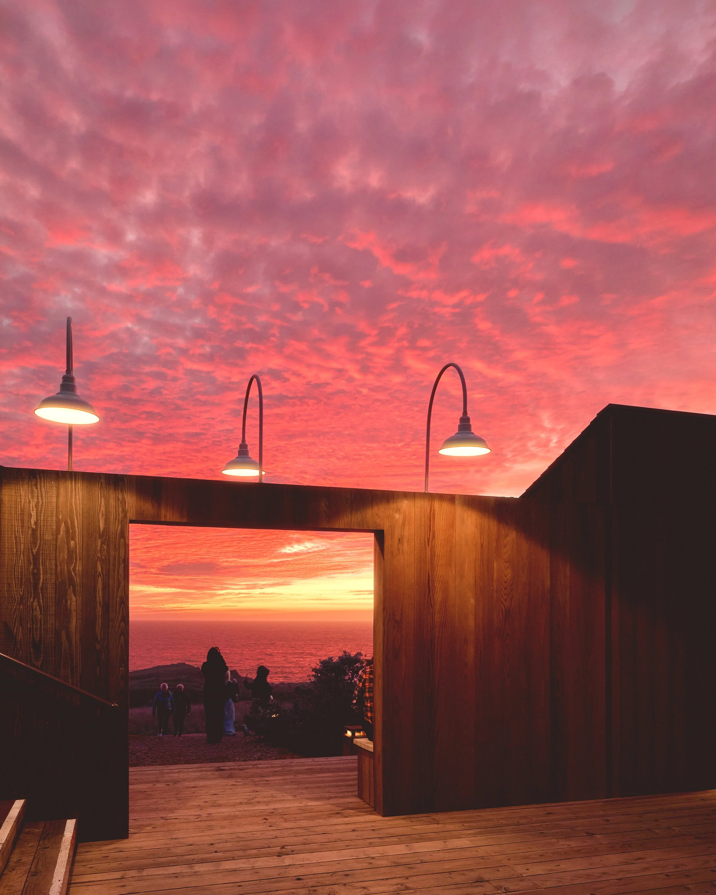 People watching a sunset from a wooden deck with three outdoor lamps, overlooking the ocean with a colorful pink and orange sky.