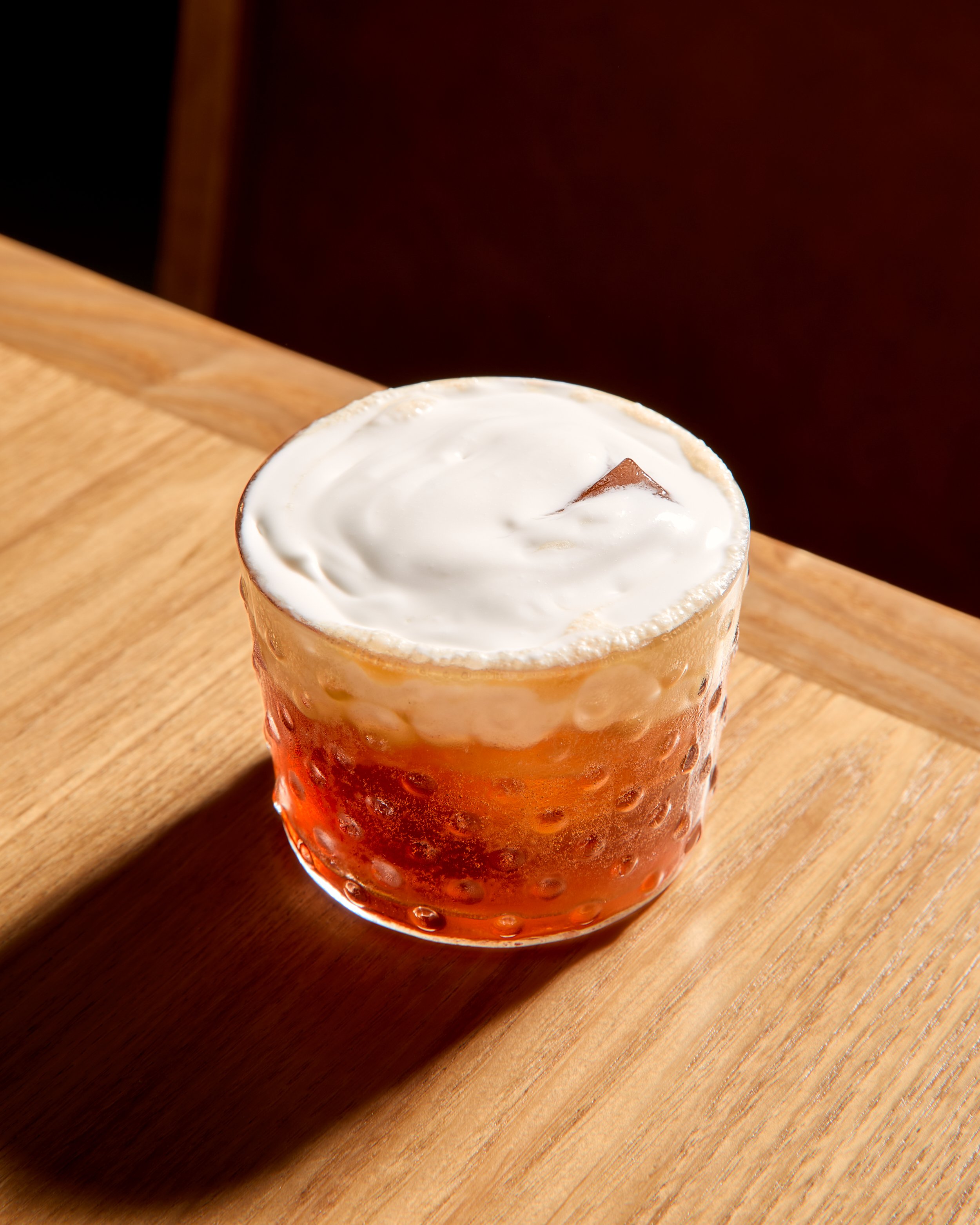 A glass of amber-colored beer topped with a layer of white foam on a wooden surface.