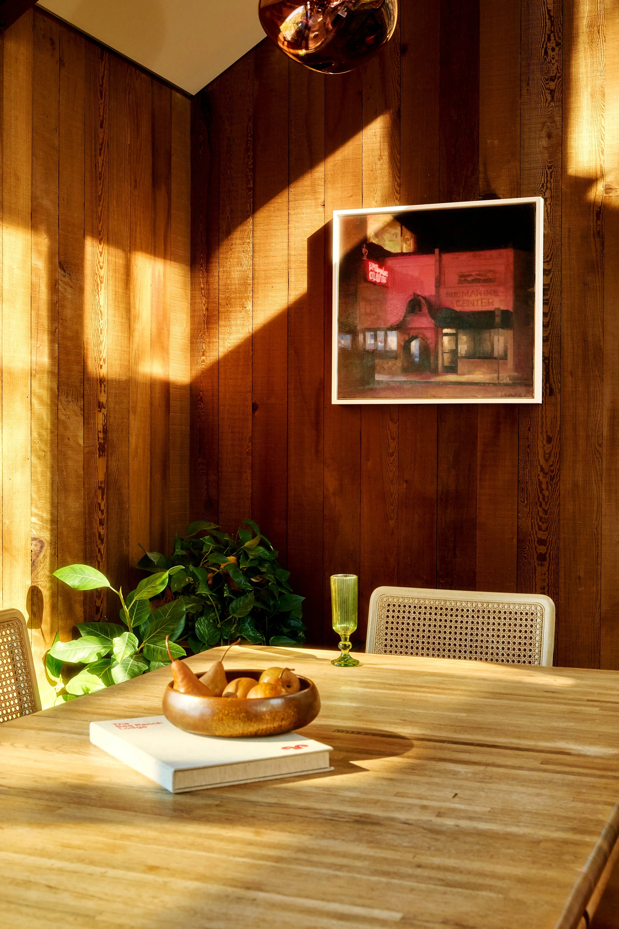 A wooden dining table with a bowl of assorted fruits, a book, and a green glass of beverage. The background features a wooden wall, a framed picture, a mustard-colored upholstered chair, and green leafy plants. Sunlight streams into the room, casting
