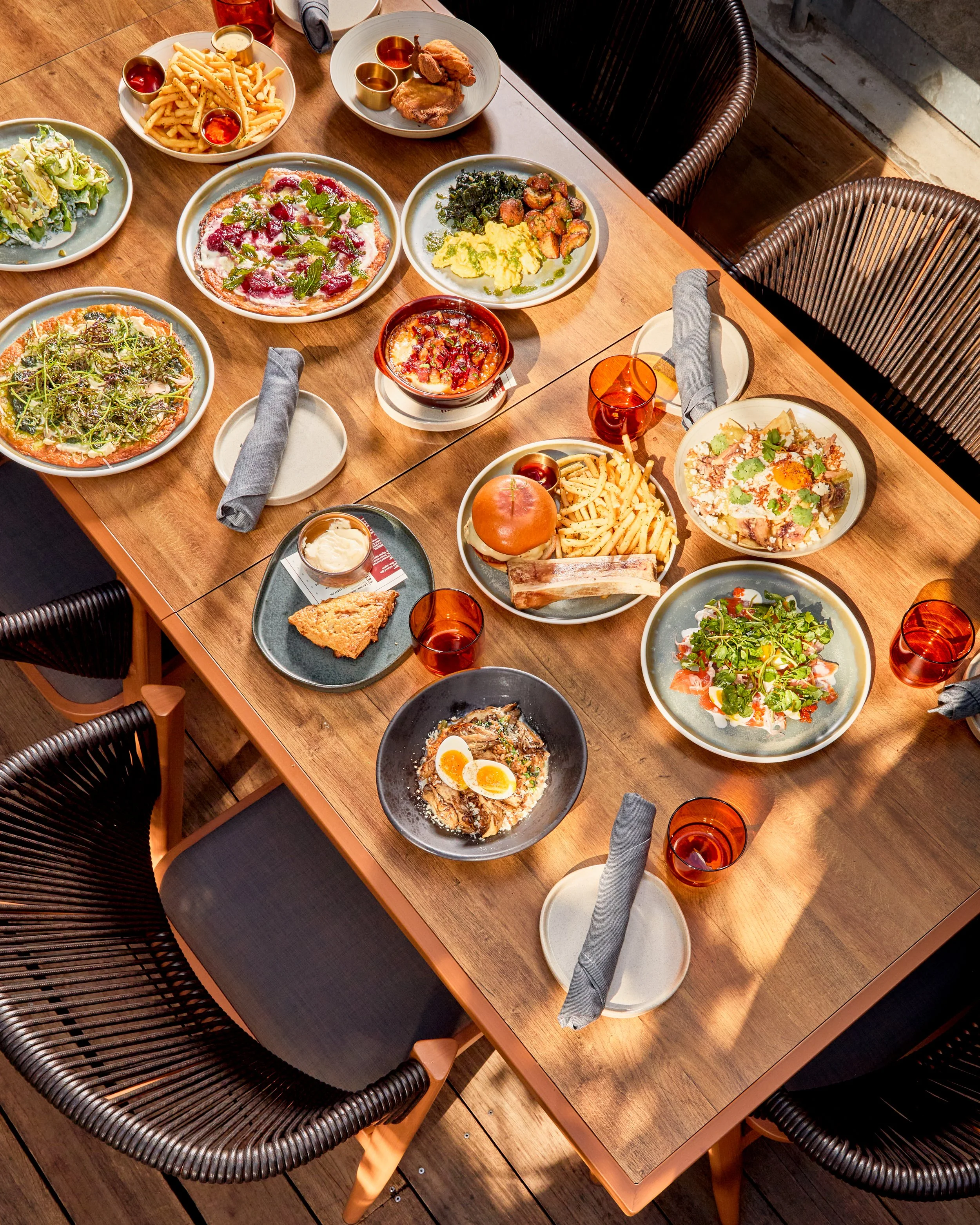 A wooden dining table set with various plates of food including pizza, fries, salad, a burger, and other dishes, surrounded by black woven chairs with gray napkins on some of the plates, and amber-colored glasses.