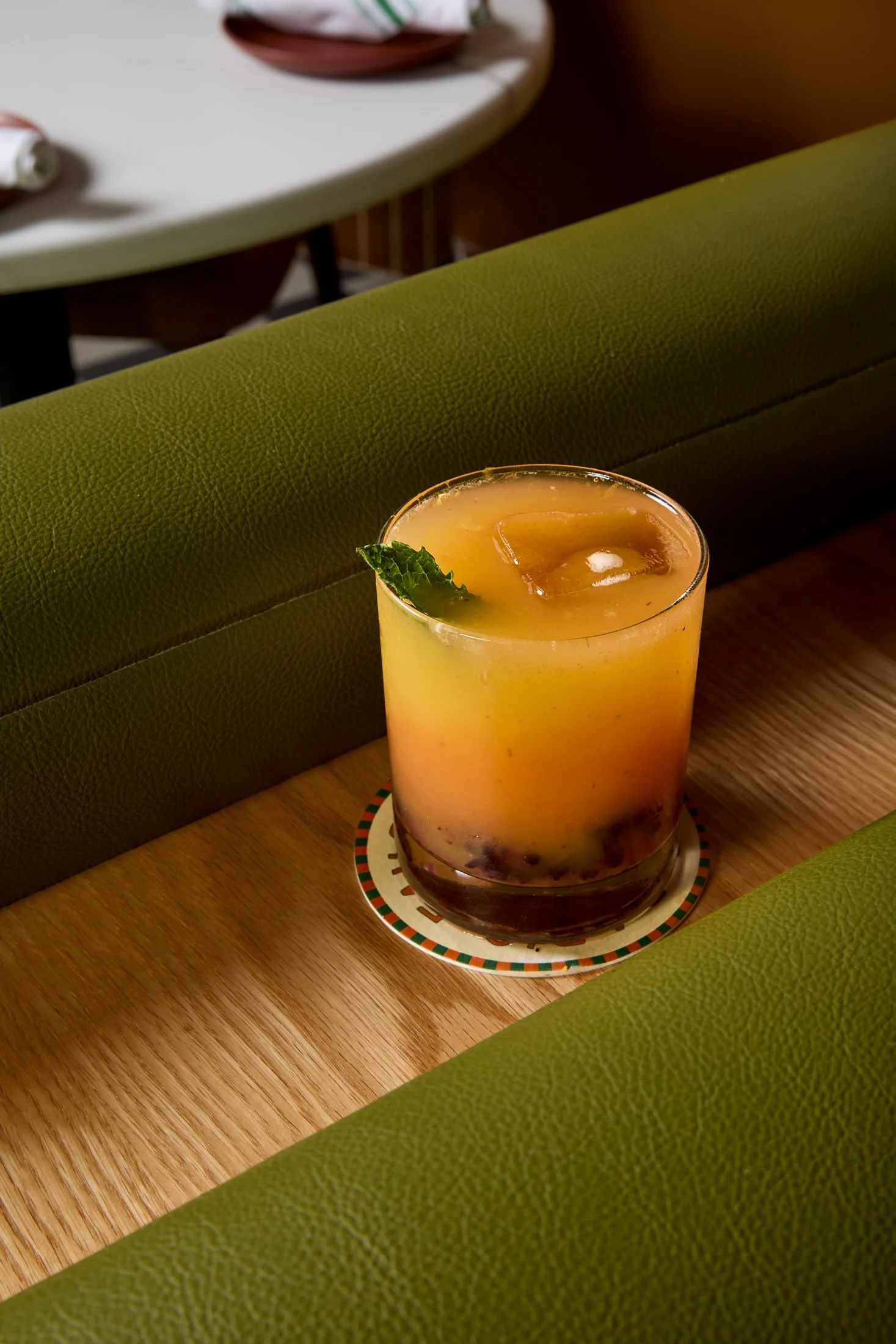 A yellow-orange cocktail with ice cubes and a fresh mint leaf garnish on a striped coaster, on a wooden table next to a green upholstered bench.