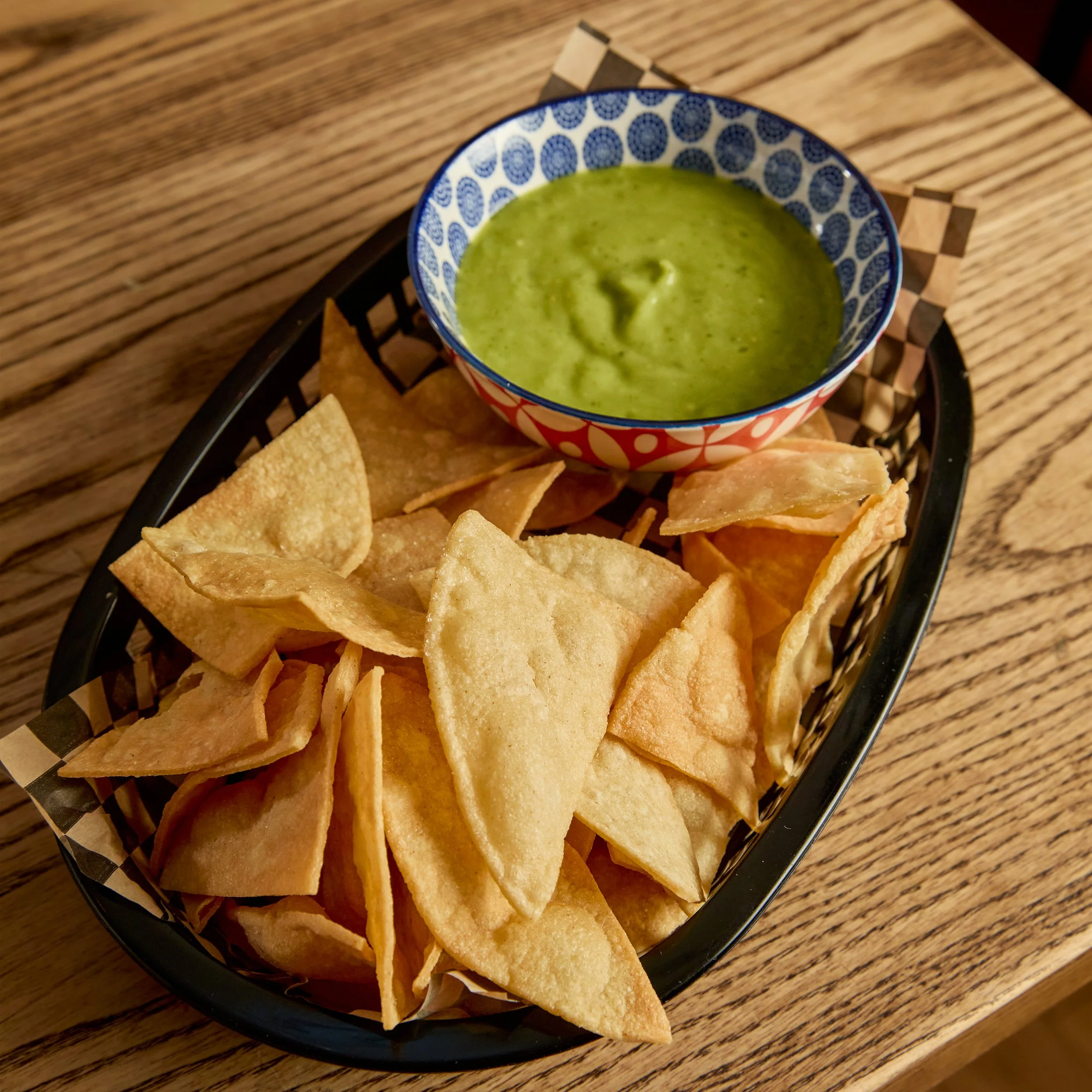 Bowl of green salsa with tortilla chips in a black basket on a wooden table.