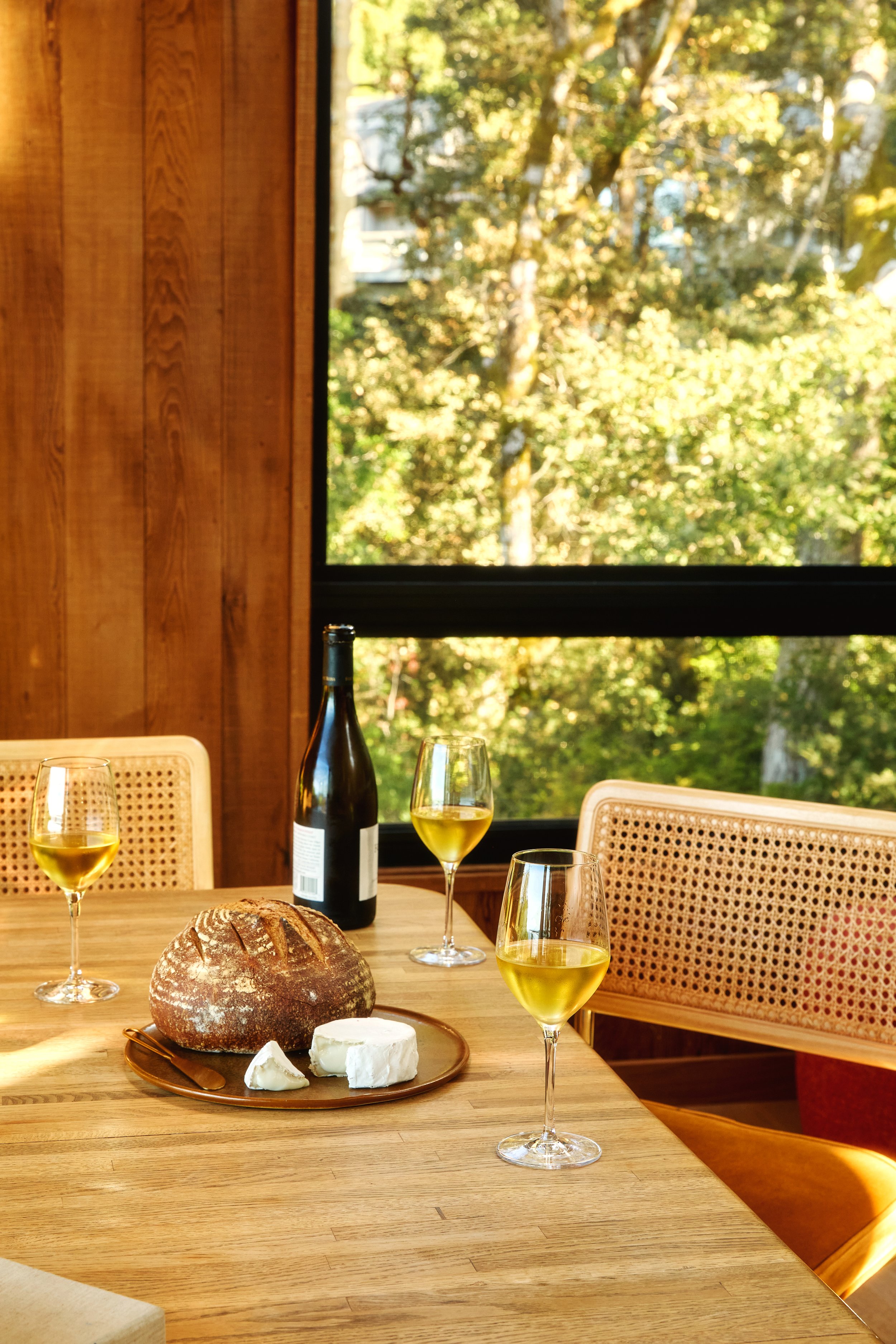 A wooden dining table set with three glasses of white wine, a loaf of bread, and a cheese platter with a round of soft cheese and a wedge, in a cozy room with large window showing green trees outside.