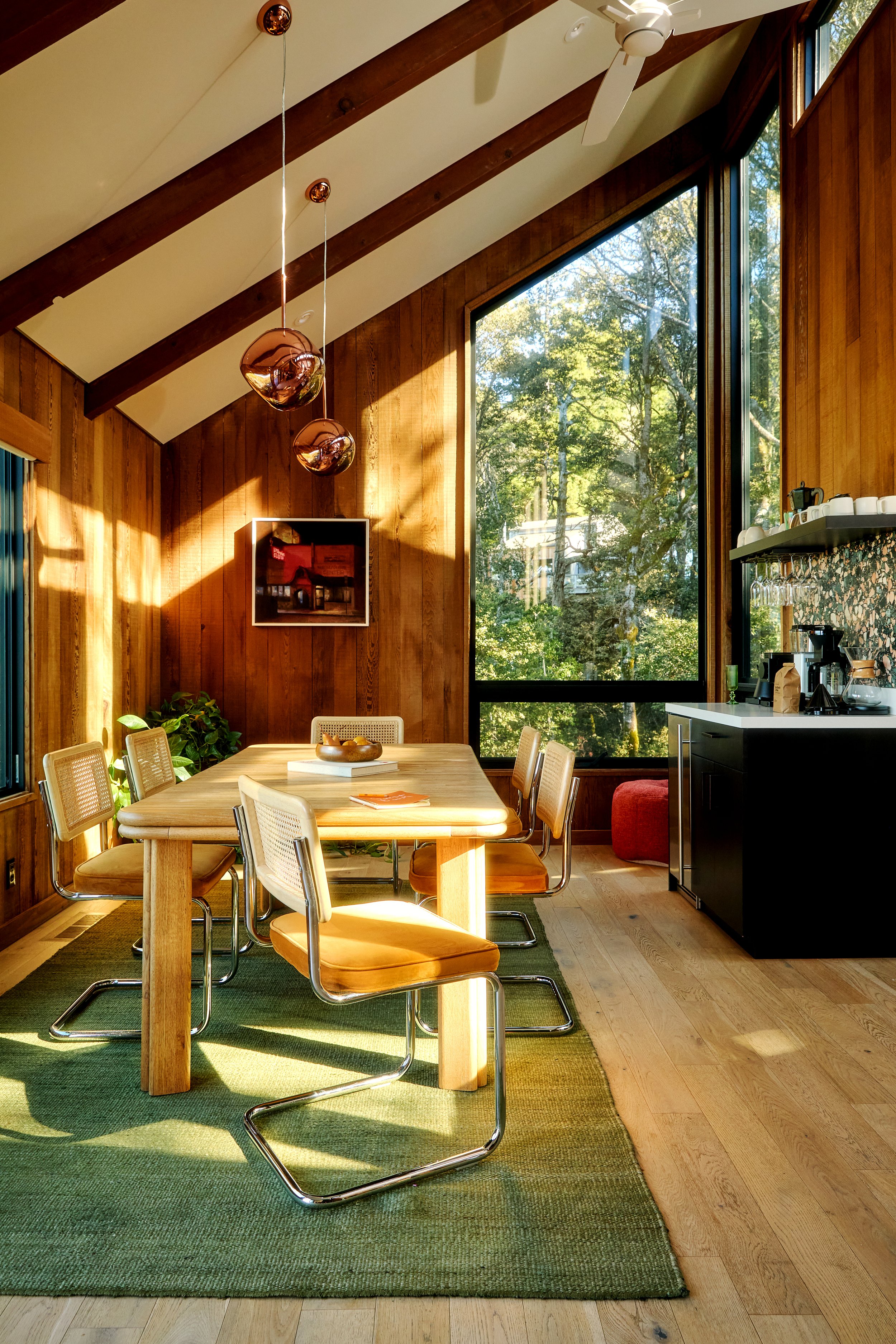 A dining area with a wooden table and six chairs, green rug, large windows showing trees outside, wooden walls, three copper pendant lights, and a small kitchen area with black cabinetry and a coffee maker.