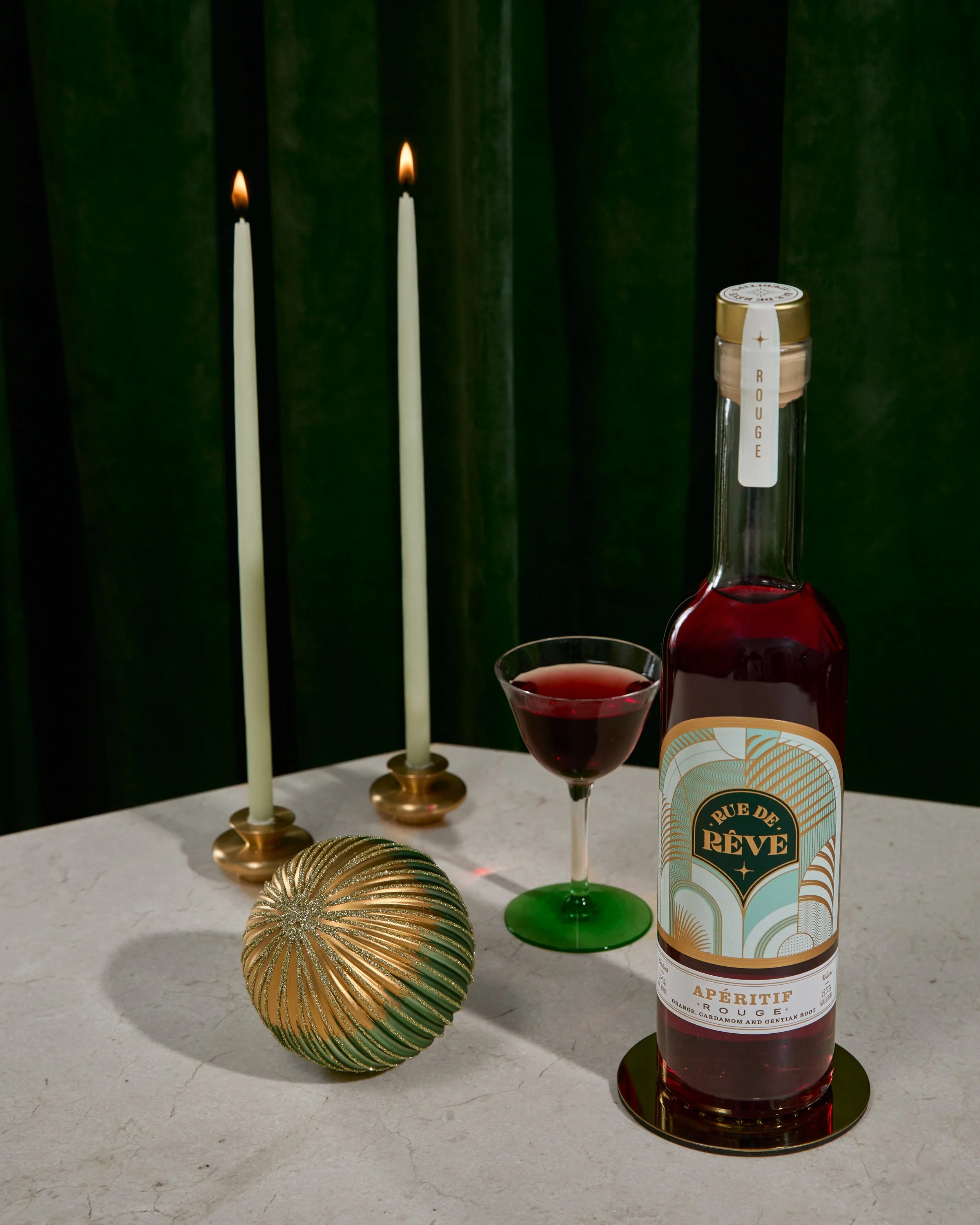 A round, gold and green decorative ornament on a white surface, two lit taper candles in brass holders, a glass of red beverage, and a bottle of Rue de Rêve Apéritif Rouge on a white surface, with a dark green curtain background.