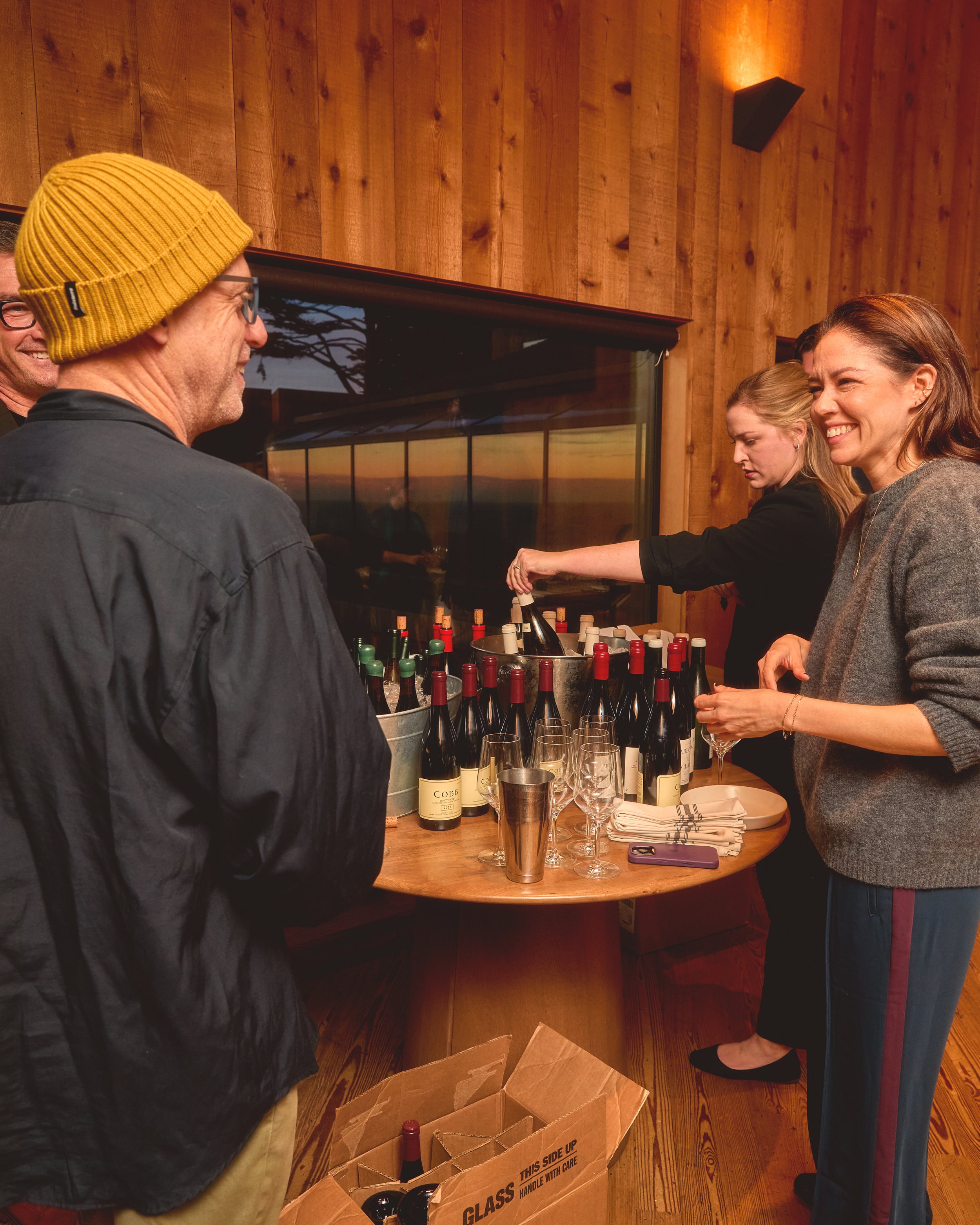 Group of people enjoying wine tasting at a wooden bar counter with bottles and glasses, with a sunset view through a window in a cozy, wood-paneled room.