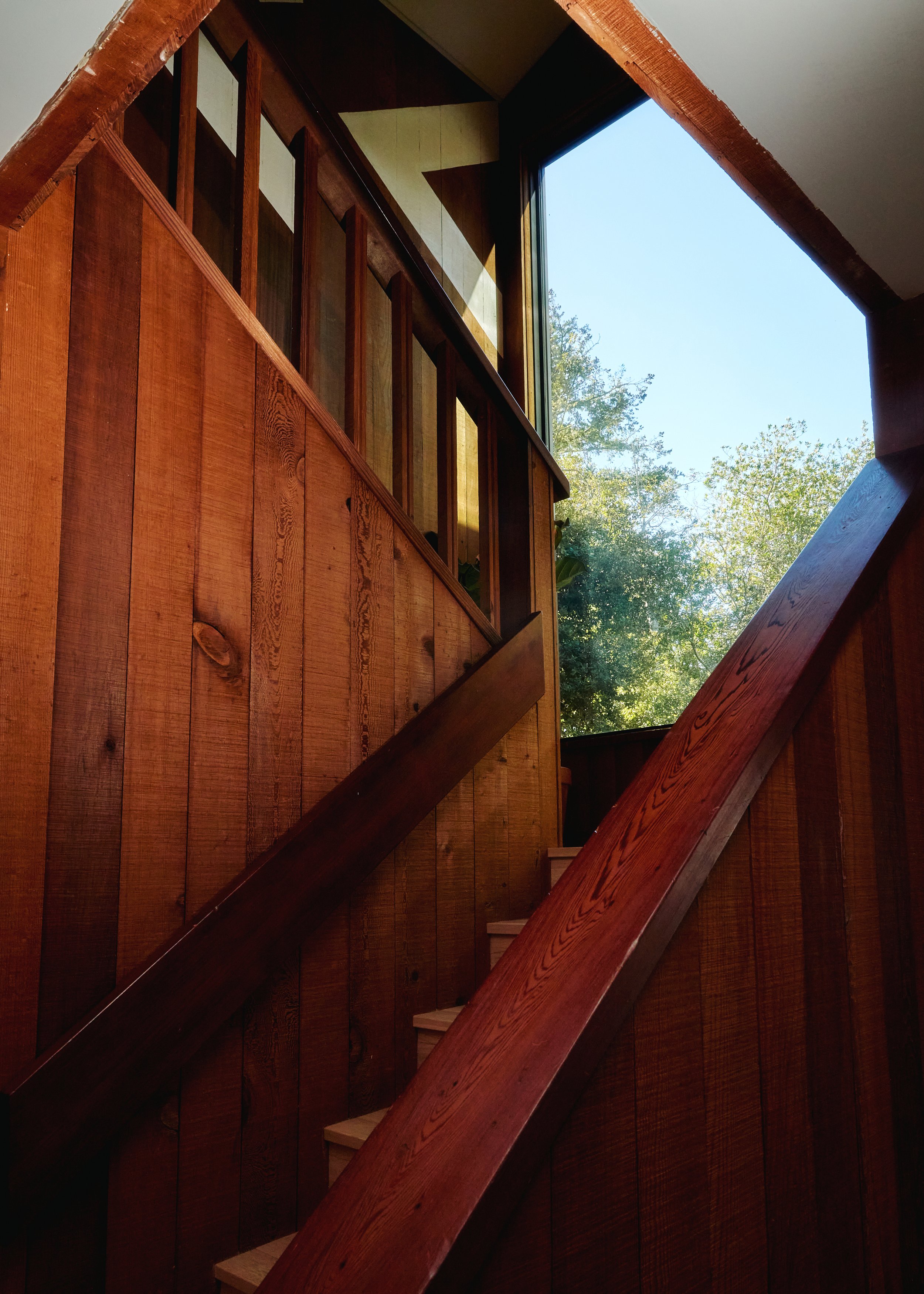 Wooden staircase with railing inside a house, opening to a large window showing trees and blue sky outside.