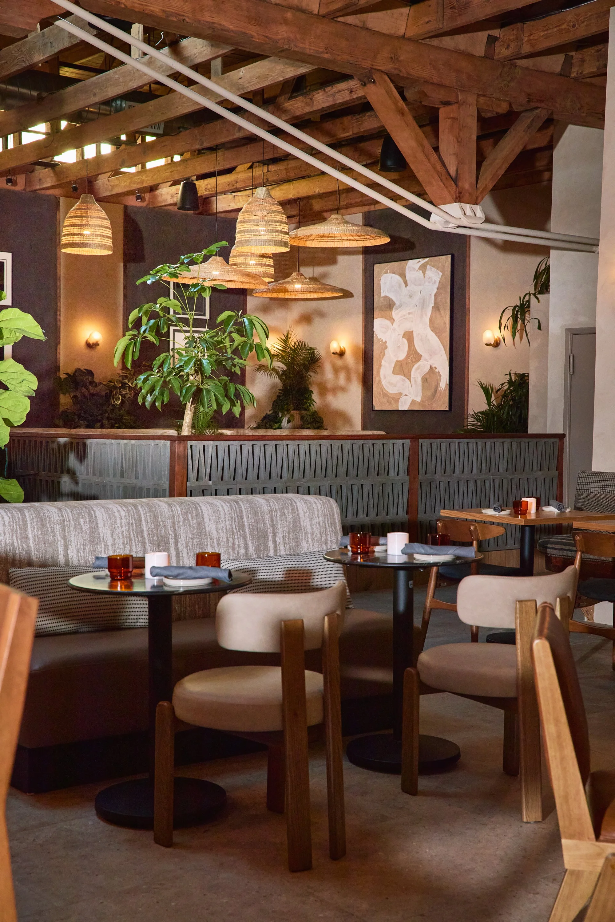 Interior of a cozy restaurant with hanging wicker light fixtures, wooden ceiling beams, and a mix of chairs and a bench seating area, decorated with plants and abstract artwork on the wall.