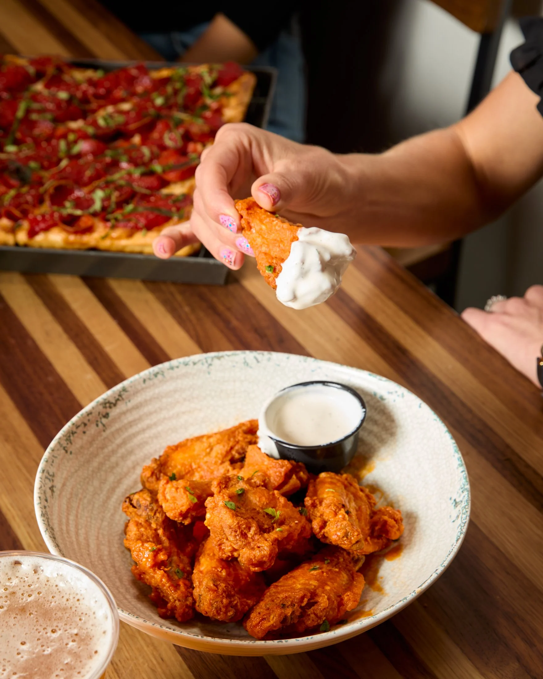 Close-up of a bowl of fried chicken with a side of dipping sauce and a tray of pizza in the background.