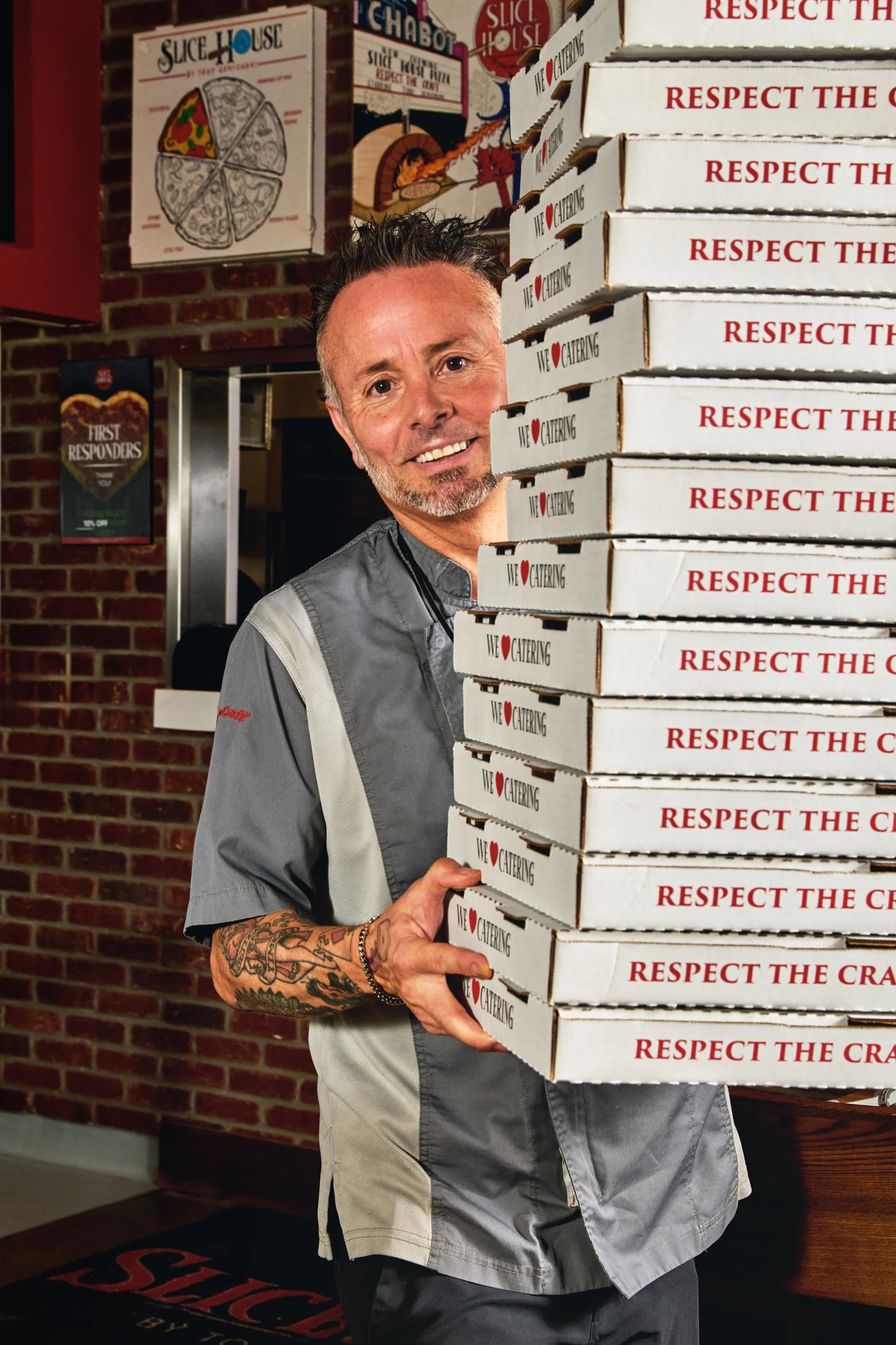 A man with tattoos and wearing a chef's uniform stands indoors holding a stack of pizza boxes labeled 'Respect the Craft' and 'We Love Catering.' Behind him are brick walls and restaurant decor including pizza-themed signs.