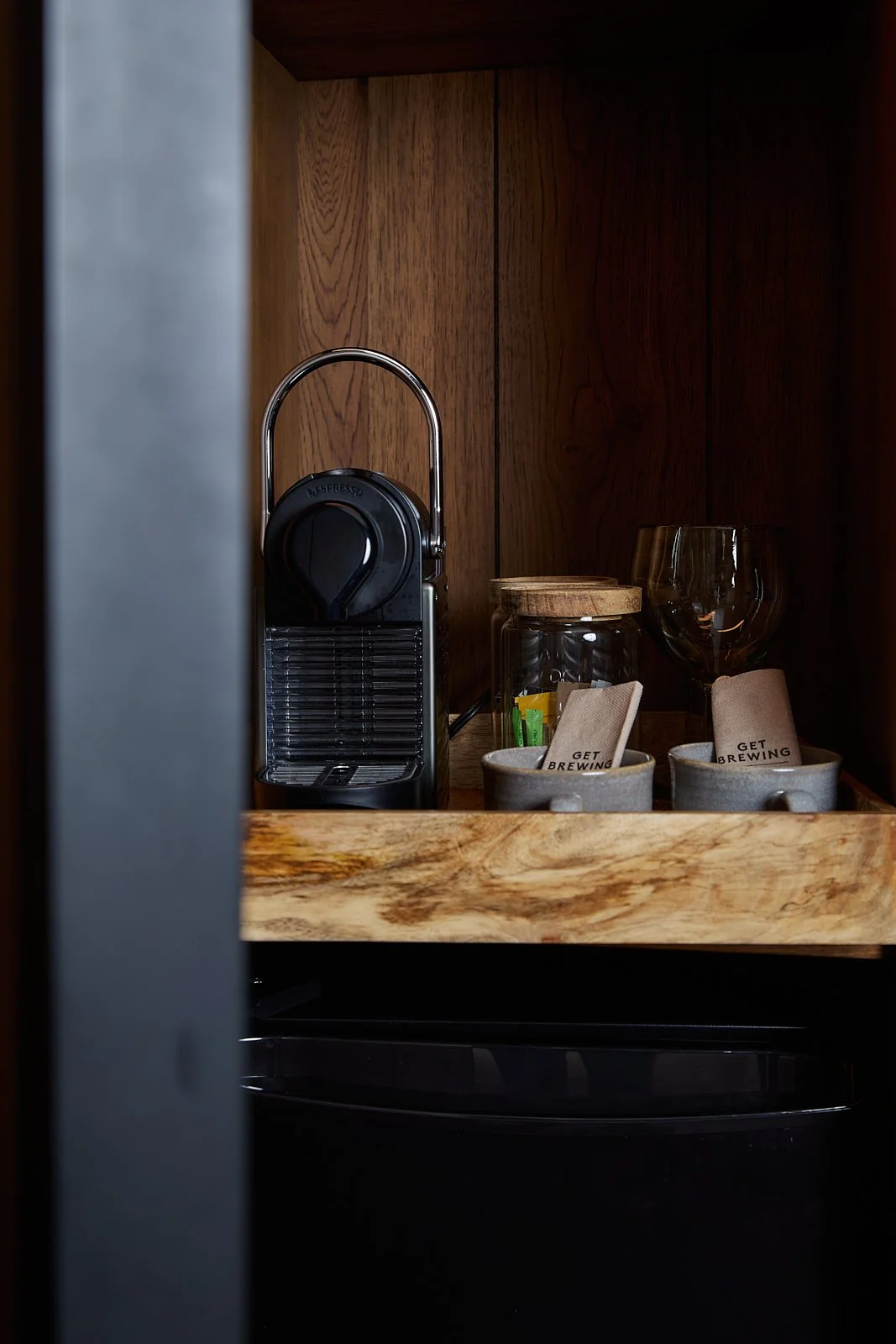 A kitchen cabinet shelf with a Nespresso coffee machine, glass jars, coffee packets, and wine glasses.