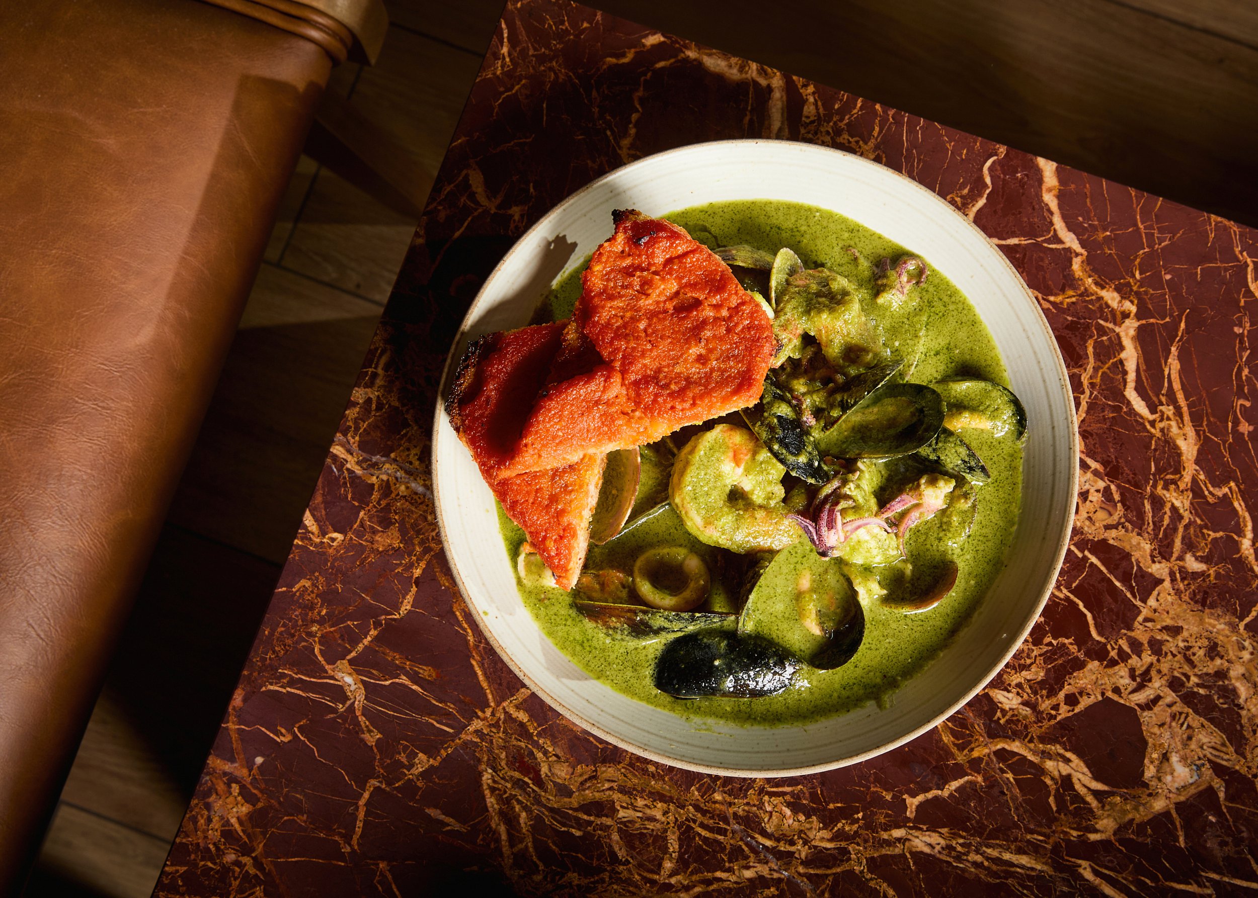 A bowl of green curry with mussels, vegetables, and a piece of fried fish on top, placed on a dark marble table.