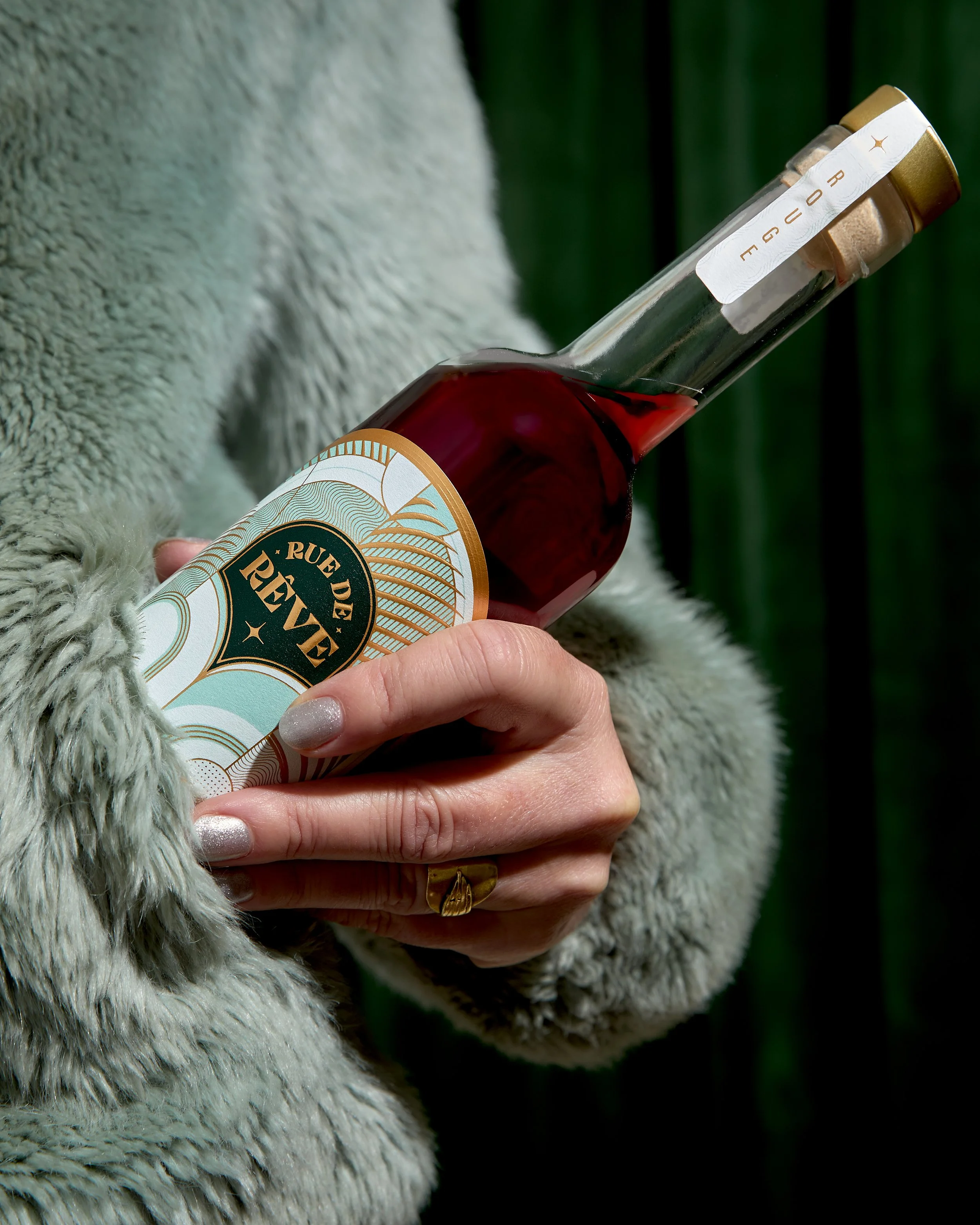 Person holding a bottle of wine with a decorative label, wearing a furry coat and a gold ring.