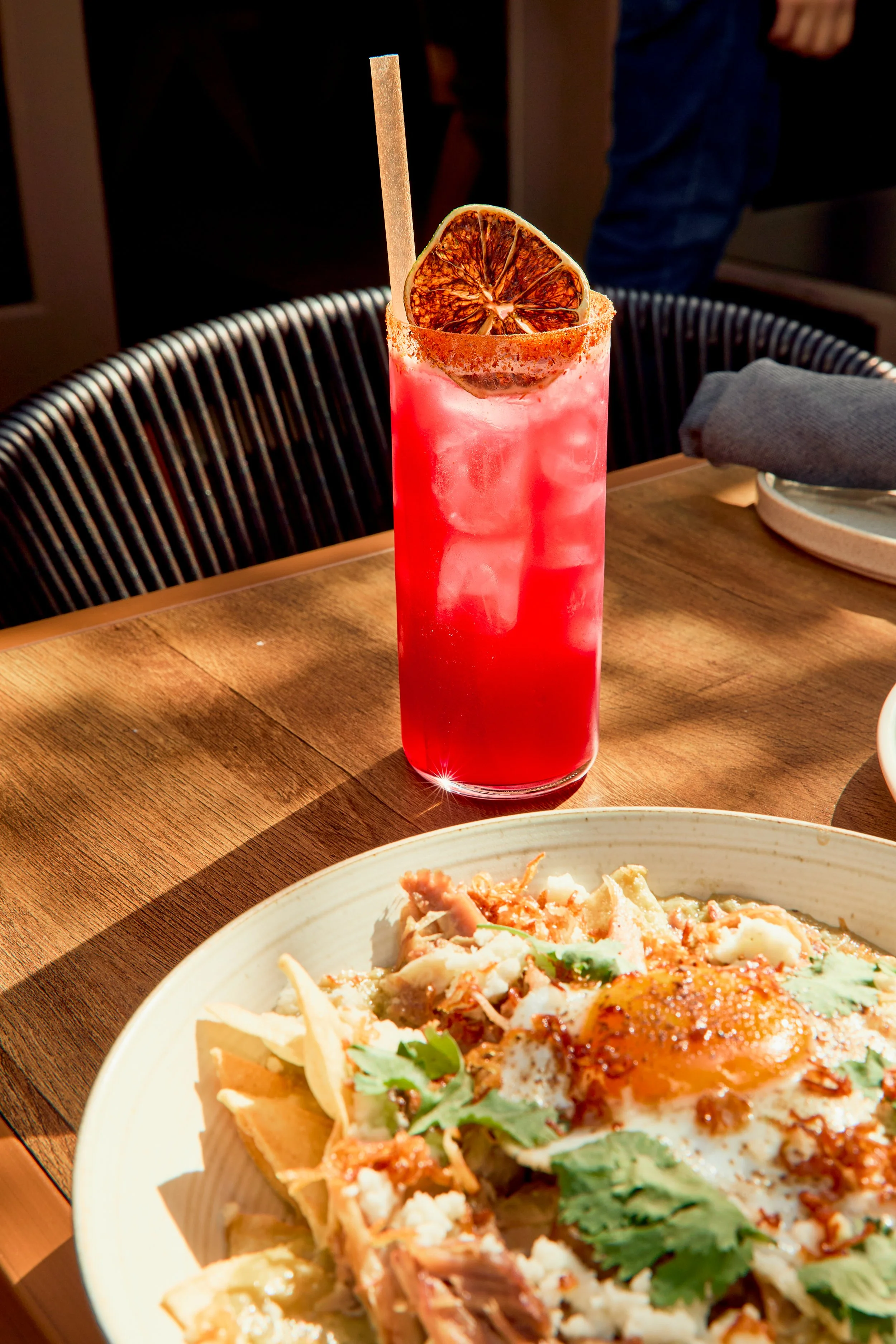 A tall glass of pink cocktail garnished with a dried orange slice and a cinnamon stick on a wooden table.