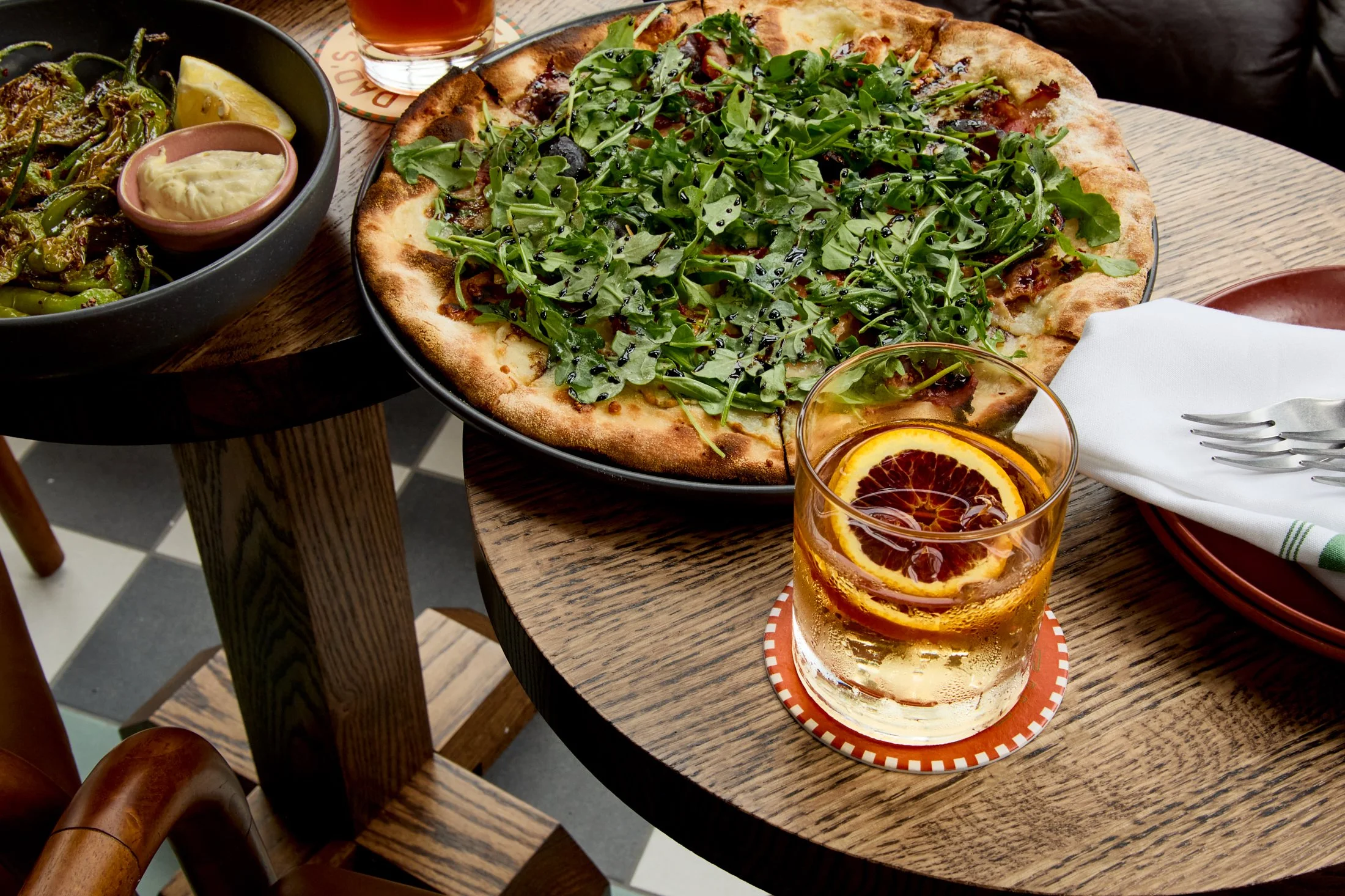 A table with a large pizza topped with arugula and black sesame seeds, a bowl of roasted green peppers with lemon wedges and sauce, a glass of iced tea with orange slices, and another dish partially visible.