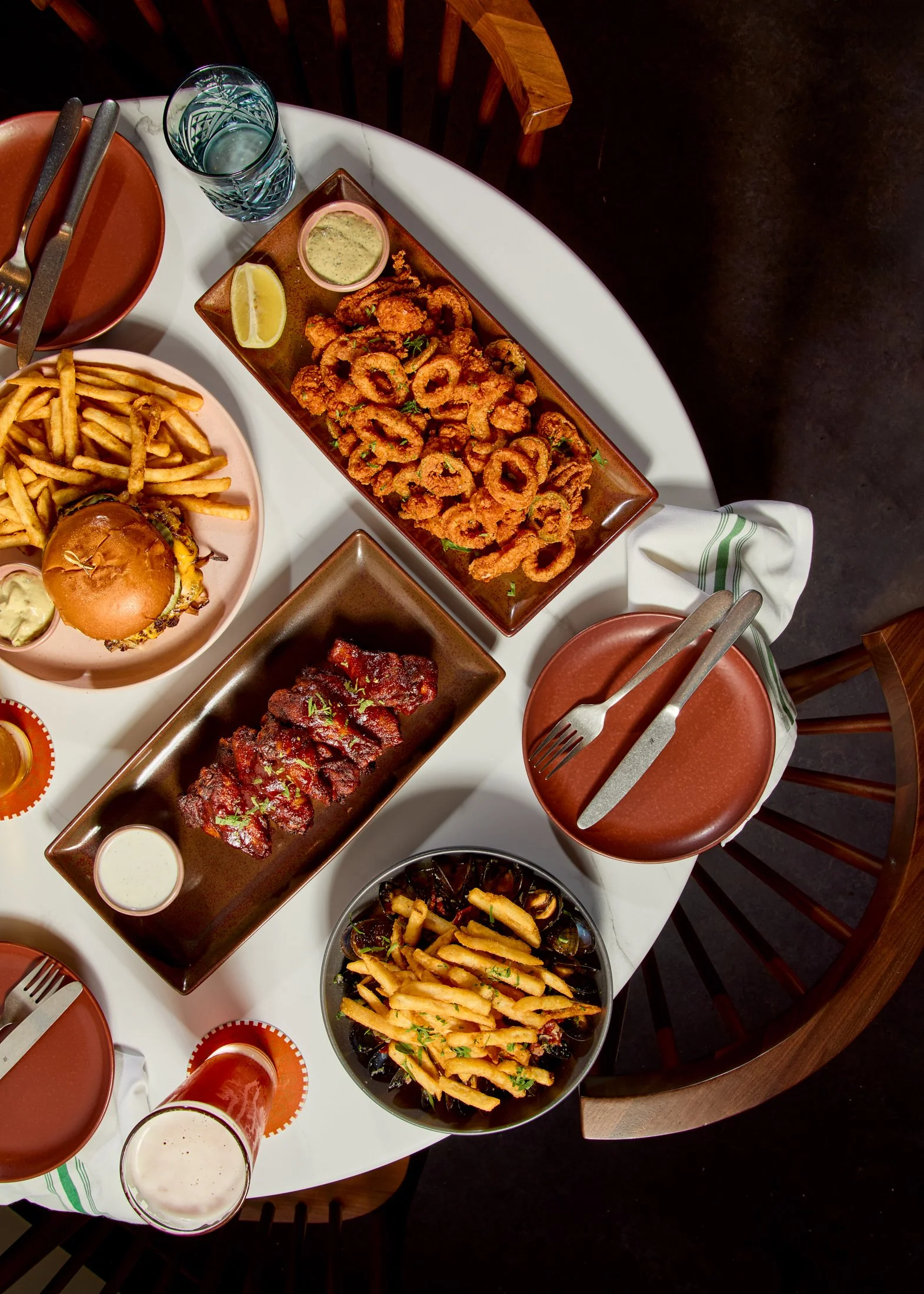 Overhead view of a dining table with various foods, including fried calamari with dipping sauce, French fries, onion rings, a burger, barbecue ribs, a glass of water, and a cup of beer, set with cutlery and plates.