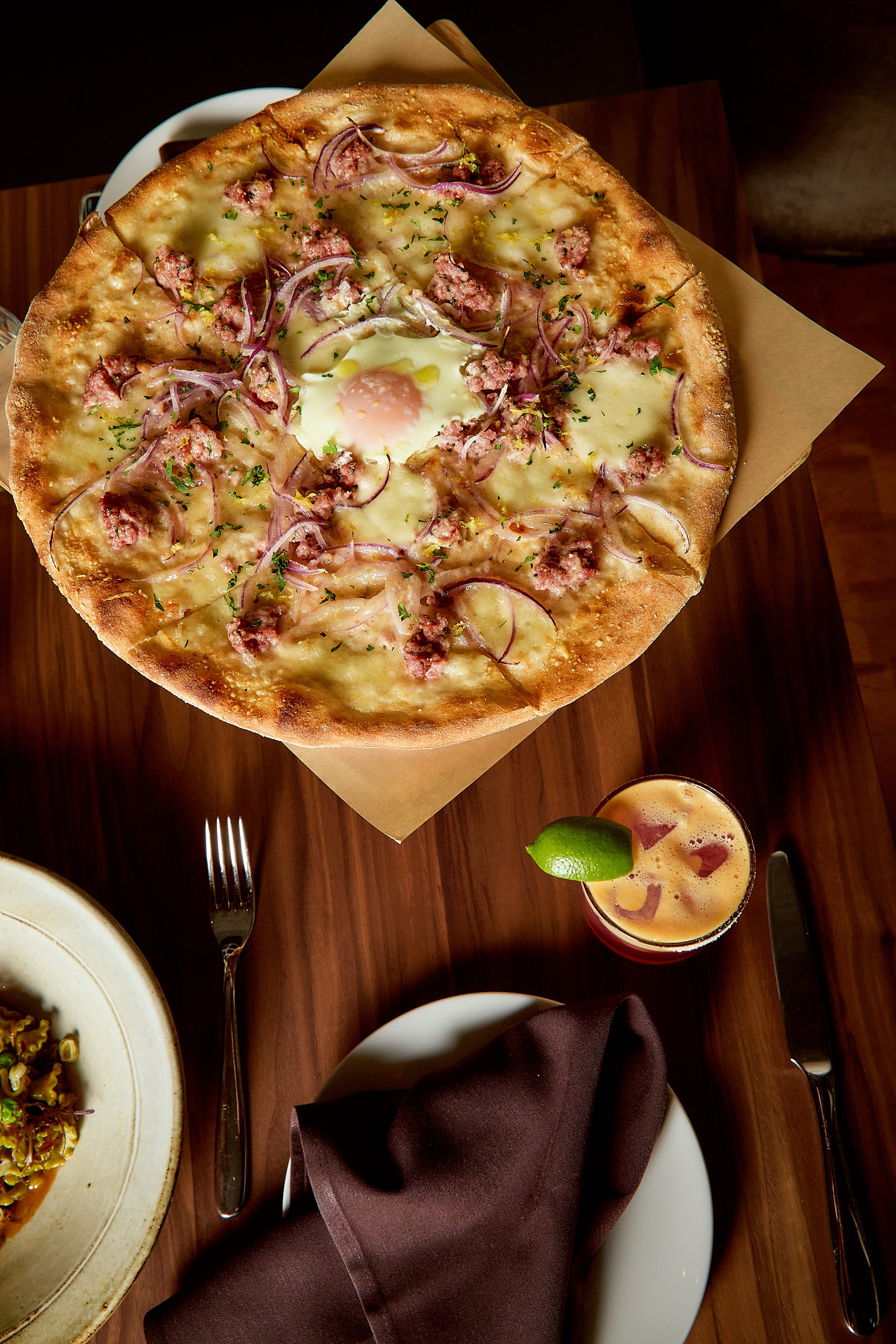 A pizza with cheese, sausage, red onions, herbs, and a fried egg in the center on a wooden table, with a cocktail garnished with lime and ice cubes nearby.