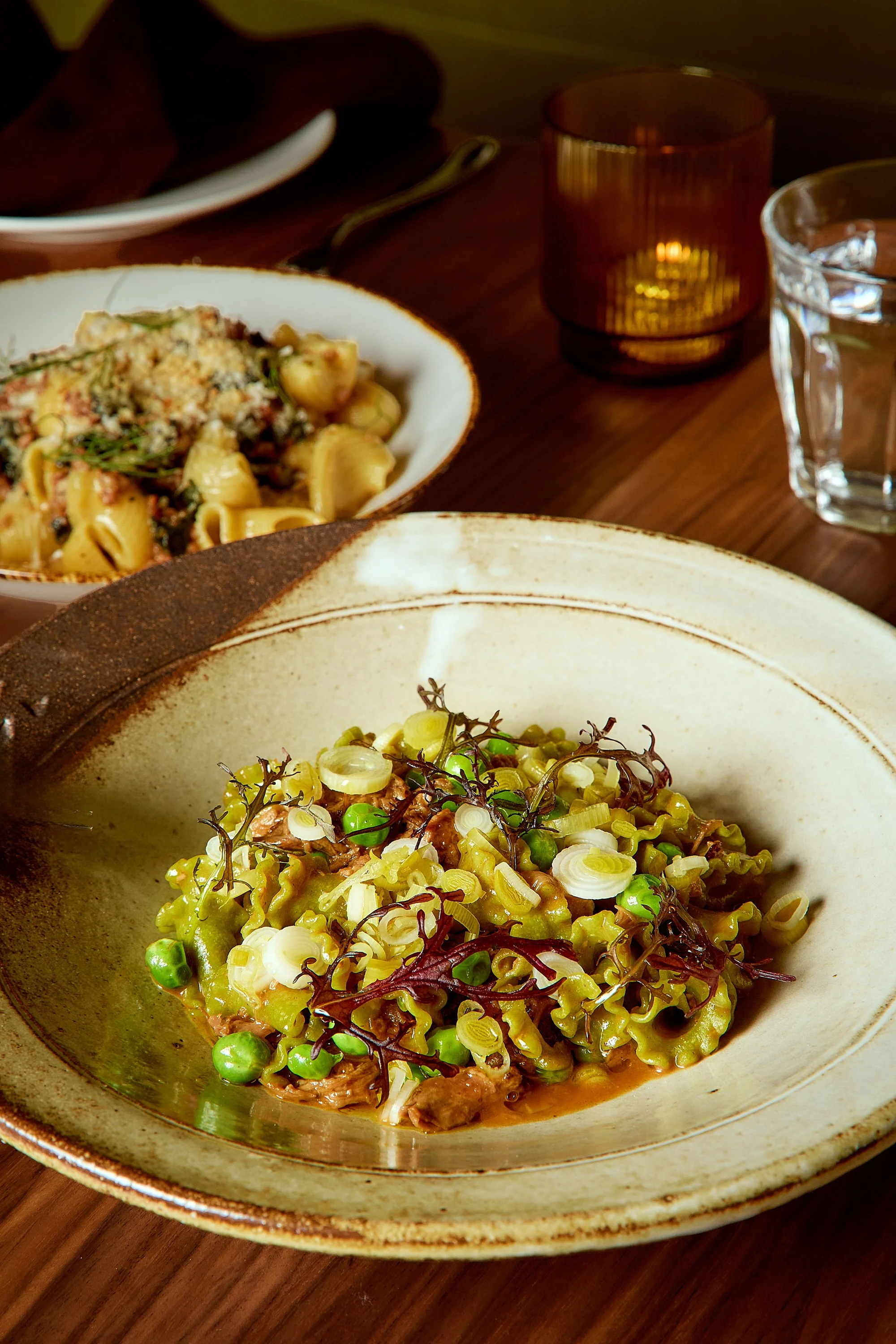 Close-up of a bowl of green pasta with green peas, herbs, and garnishes, served on a rustic ceramic plate on a wooden table.