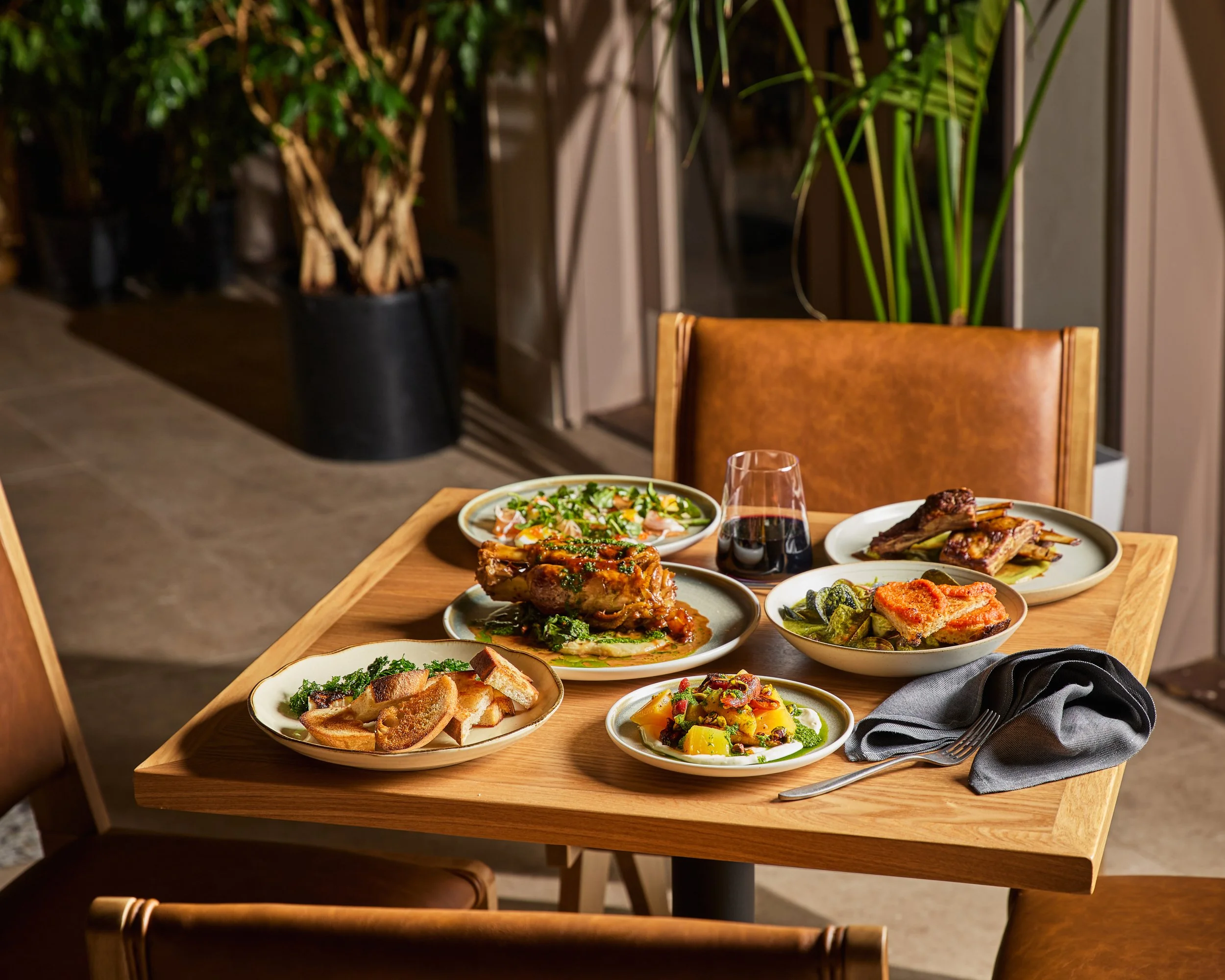 A wooden dining table set with several plates of food, including roasted chicken, vegetables, bread, and salad, along with a glass of red wine, in a well-lit restaurant dining area.