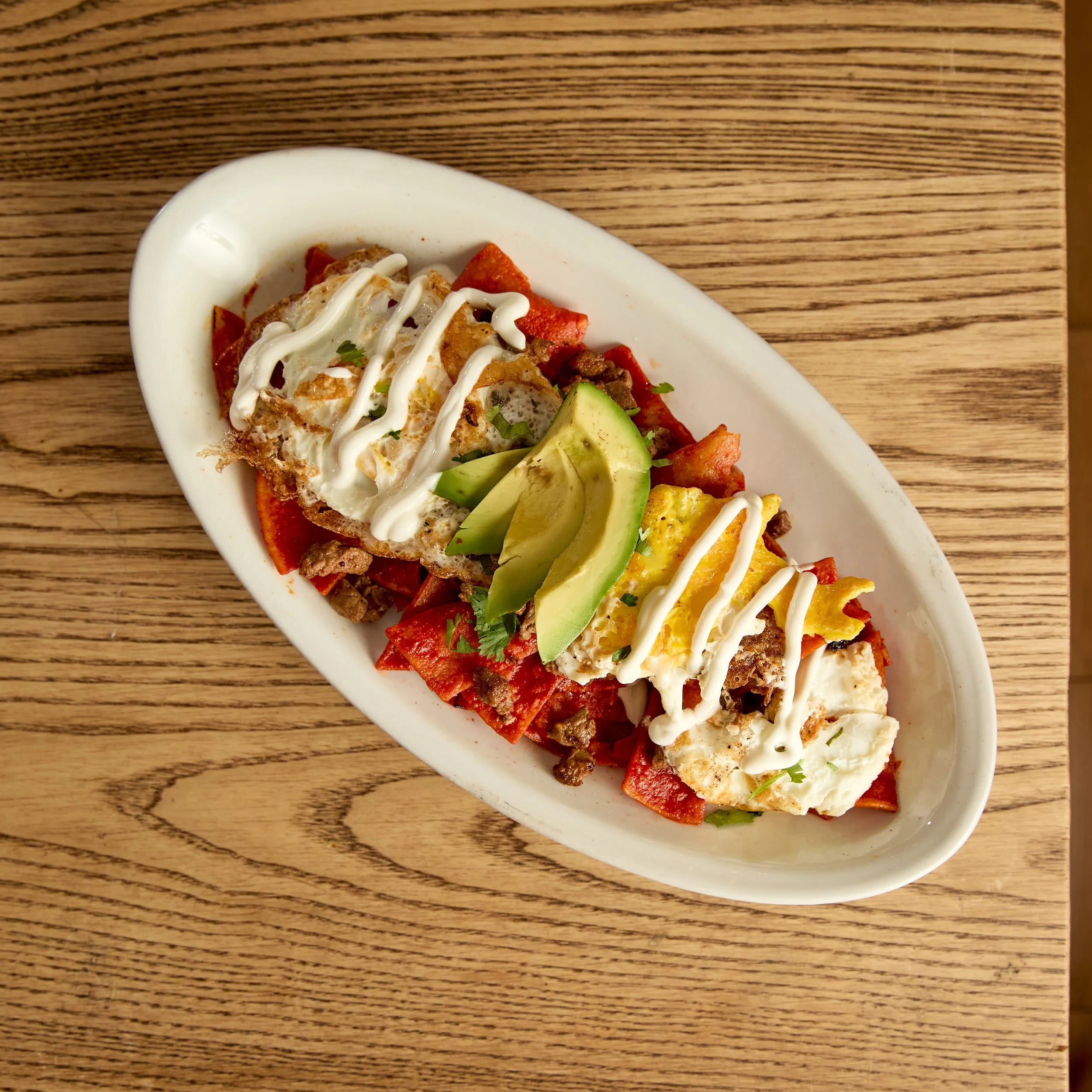 Overhead view of a serving of chilaquiles with fried eggs, avocado slices, cotija cheese, and drizzled sour cream on a white oval dish on a wooden table.