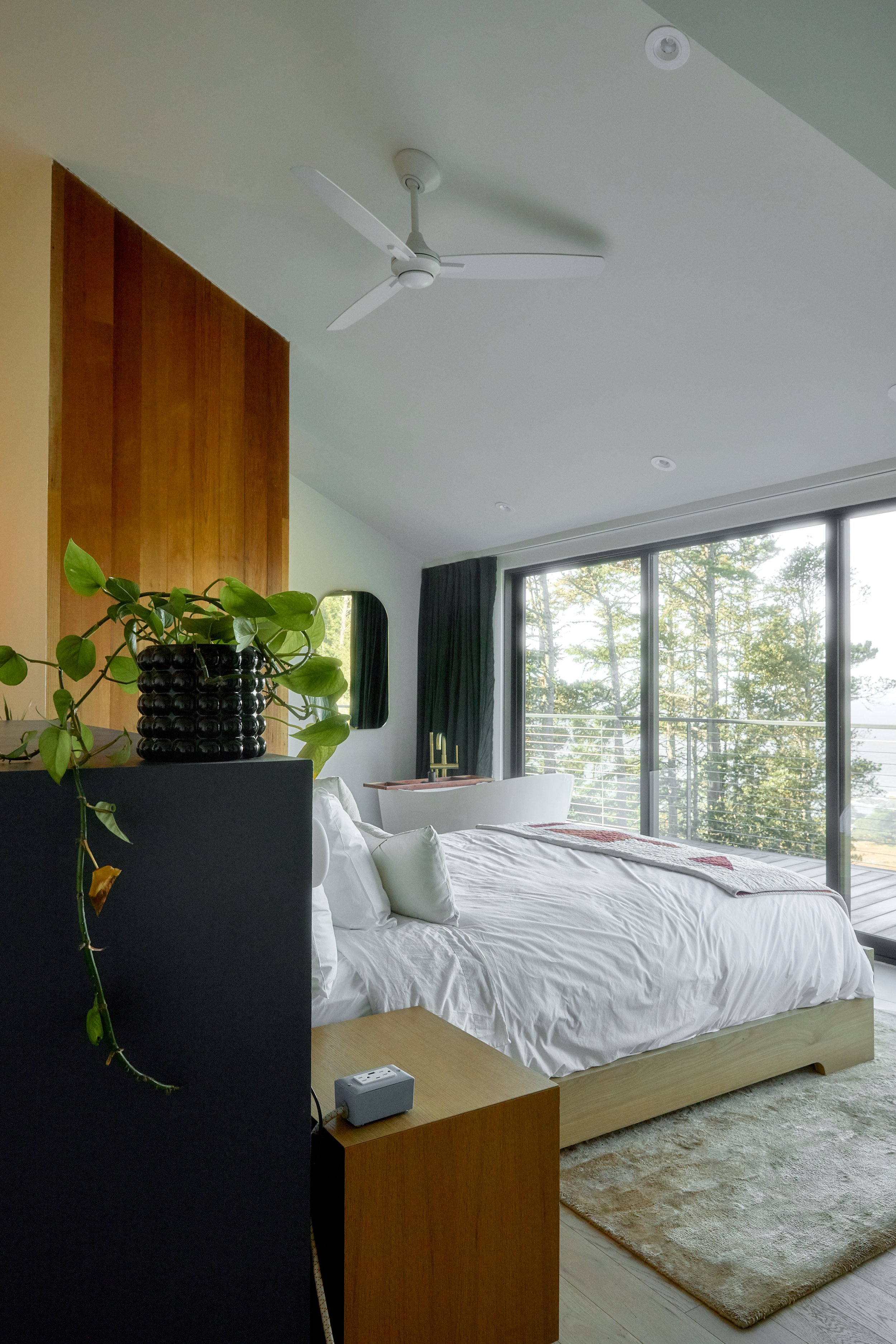 Modern bedroom with a large bed, white bedding, a bedside table with a speaker, a green plant, a window with a view of trees, and a ceiling fan.