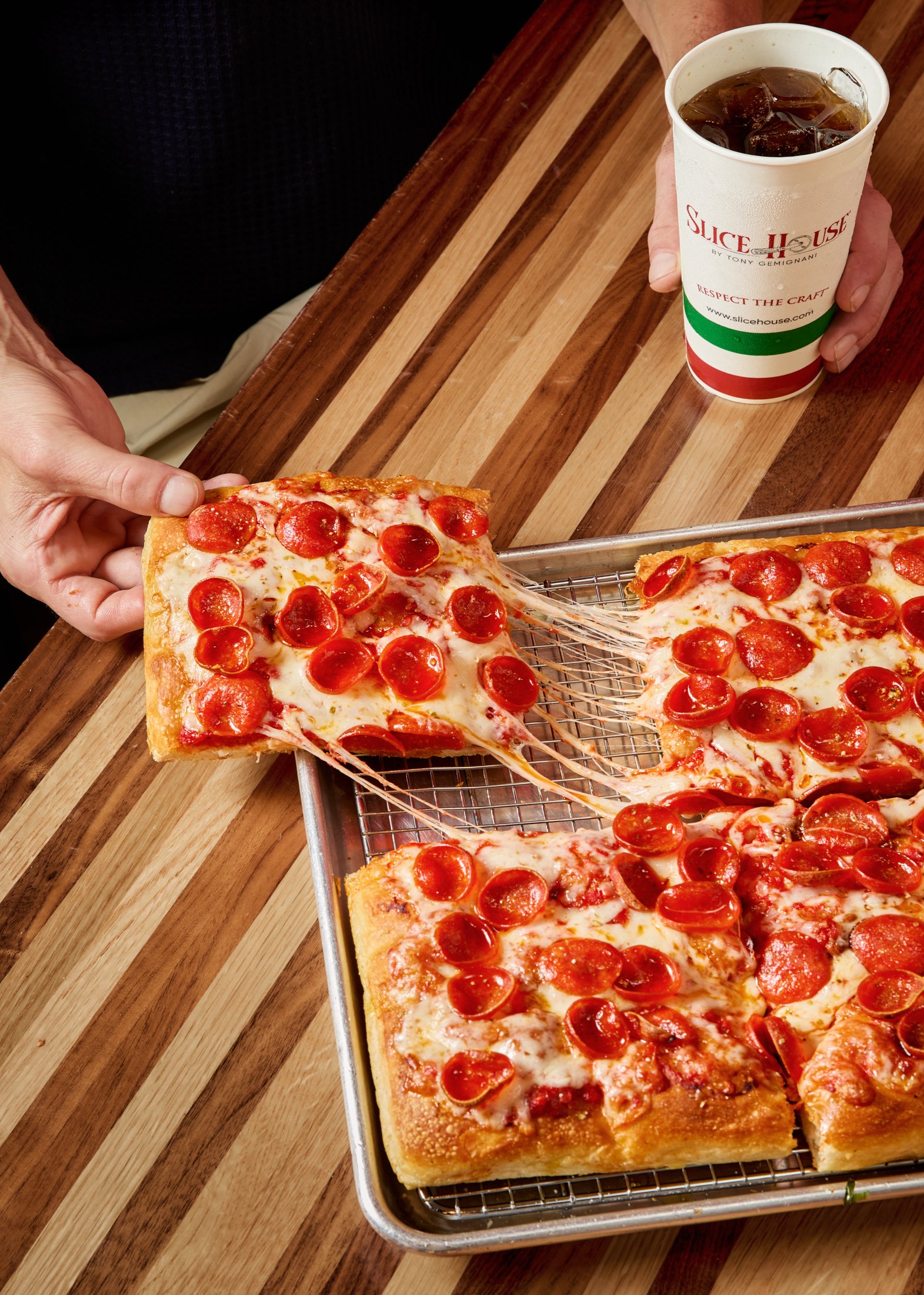A person holding a slice of pepperoni pizza next to a tray of pizza slices on a wire rack, with a cup of iced soda on a wooden table.