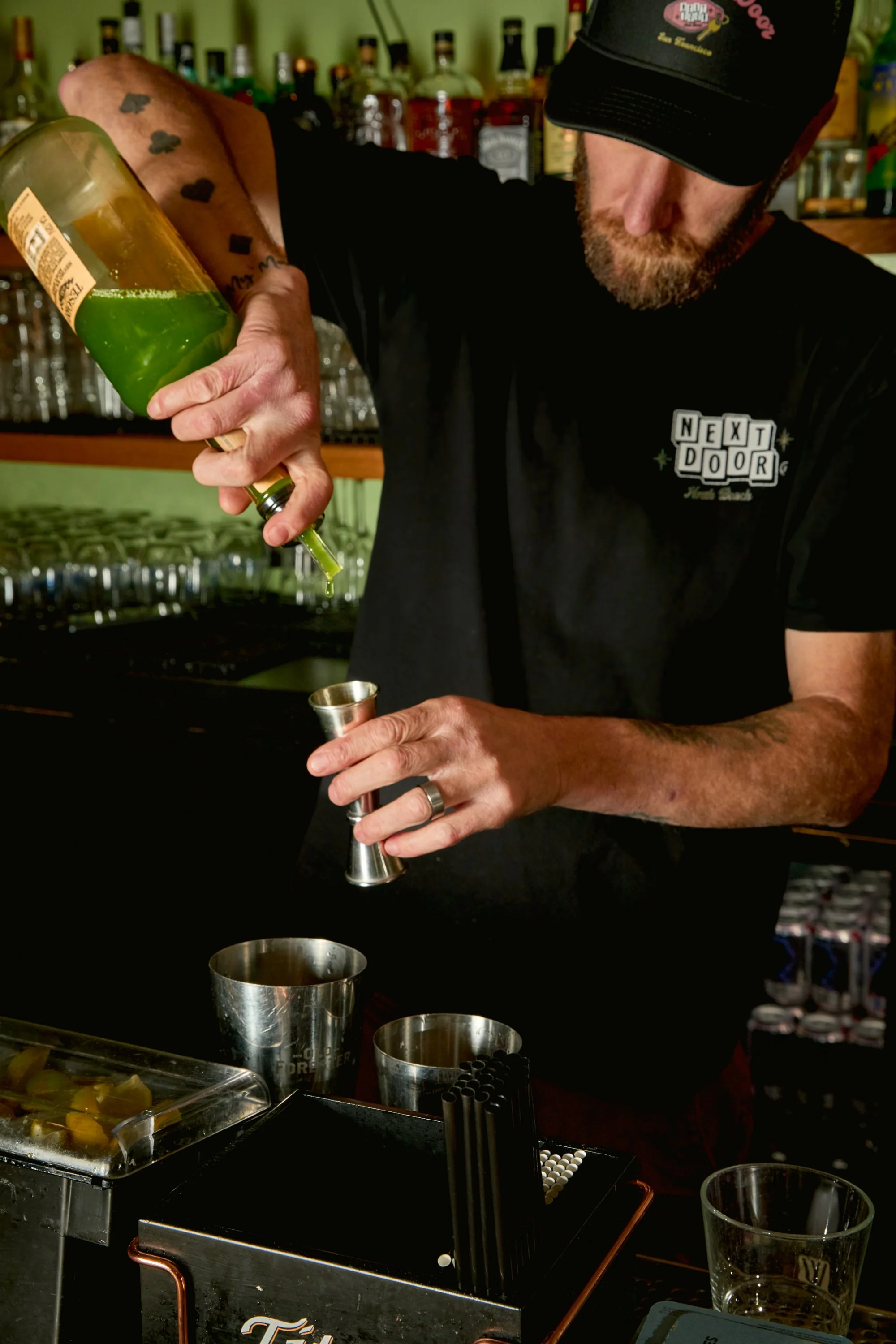 A bartender pouring green liquid into a jigger at a bar with bottles of alcohol in the background.