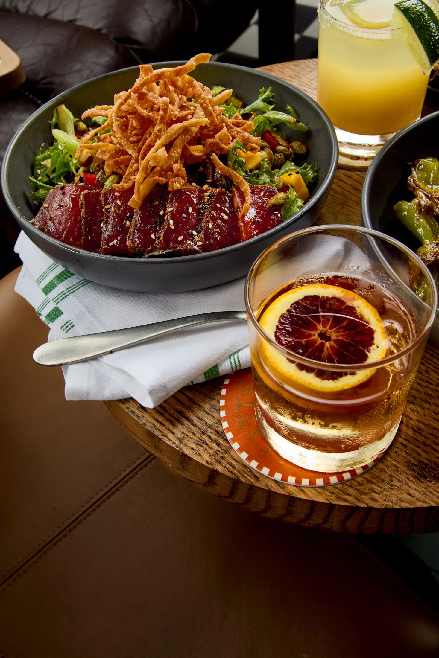 A black bowl filled with a salad containing mixed greens, sliced red peppers, and crispy fried strips, topped with slices of raw tuna sprinkled with sesame seeds. Next to the bowl is a glass of iced tea with lemon and orange slices. Part of another b