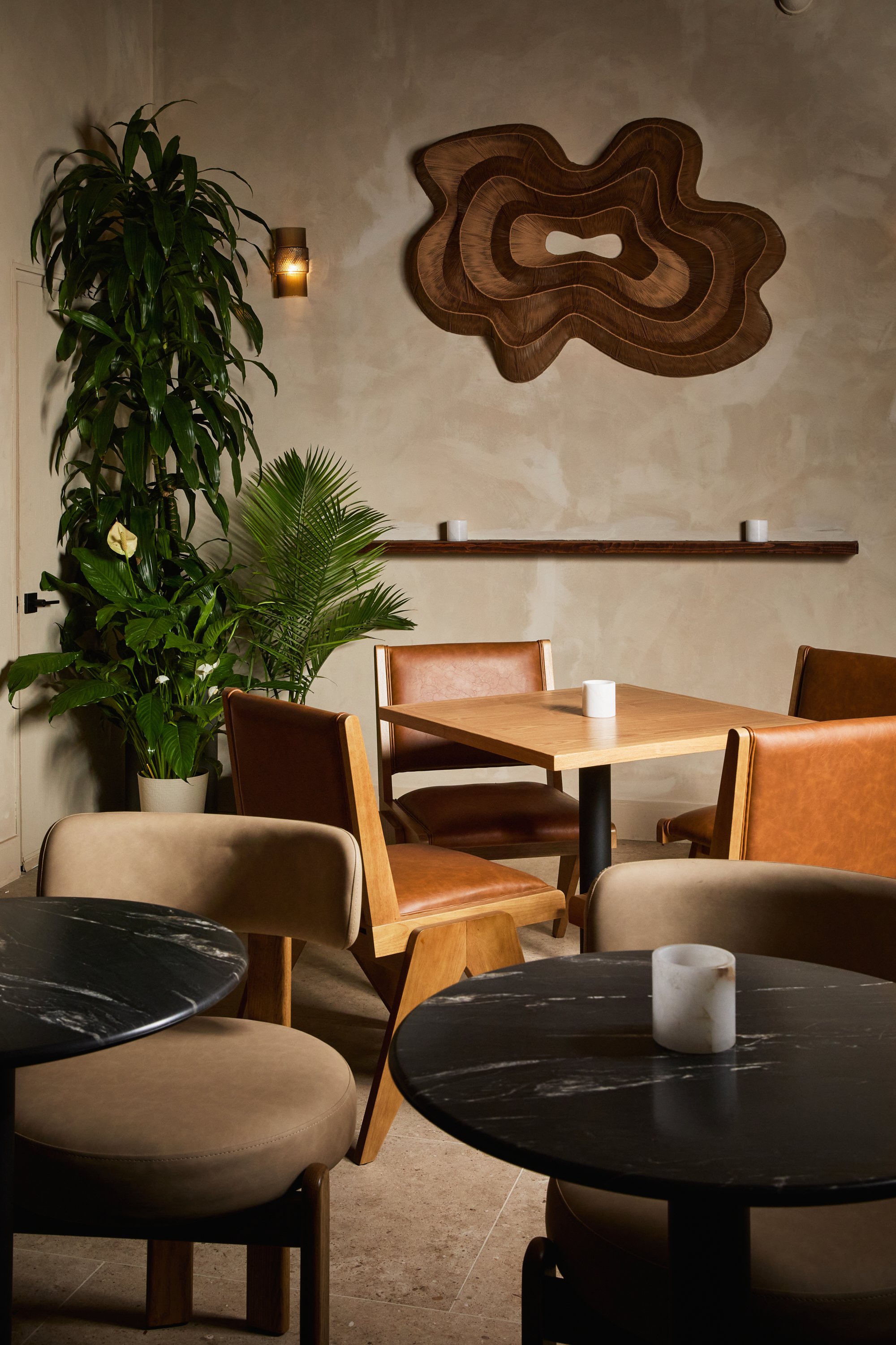 A cozy interior of a café or restaurant with a wooden table, leather and fabric chairs, a black marble table with a candle, green plants, a wave-shaped wall art, and candles on a shelf.