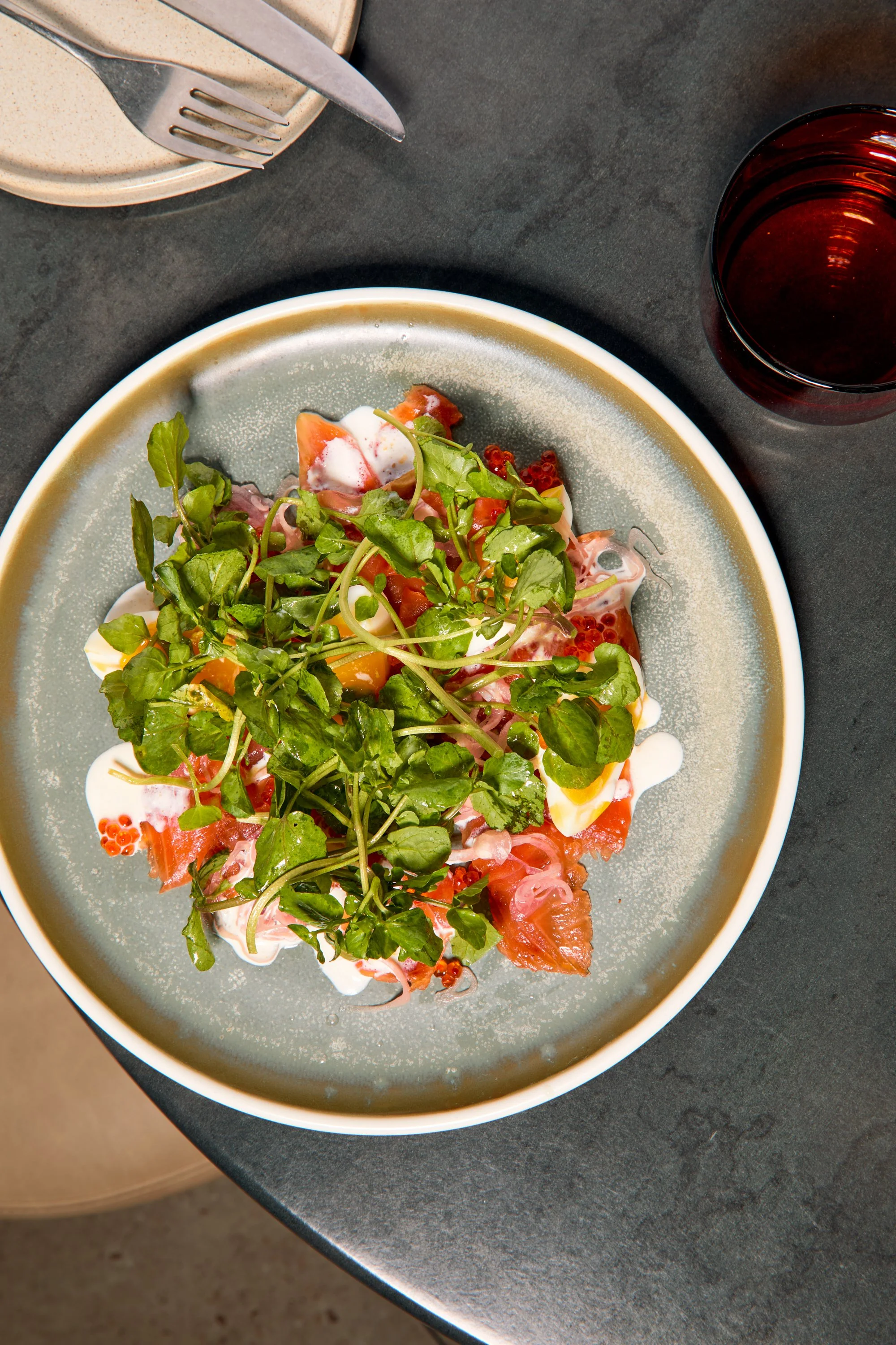 A plate of smoked salmon topped with fresh greens and a creamy sauce, set on a dark gray table next to a glass of dark beverage.