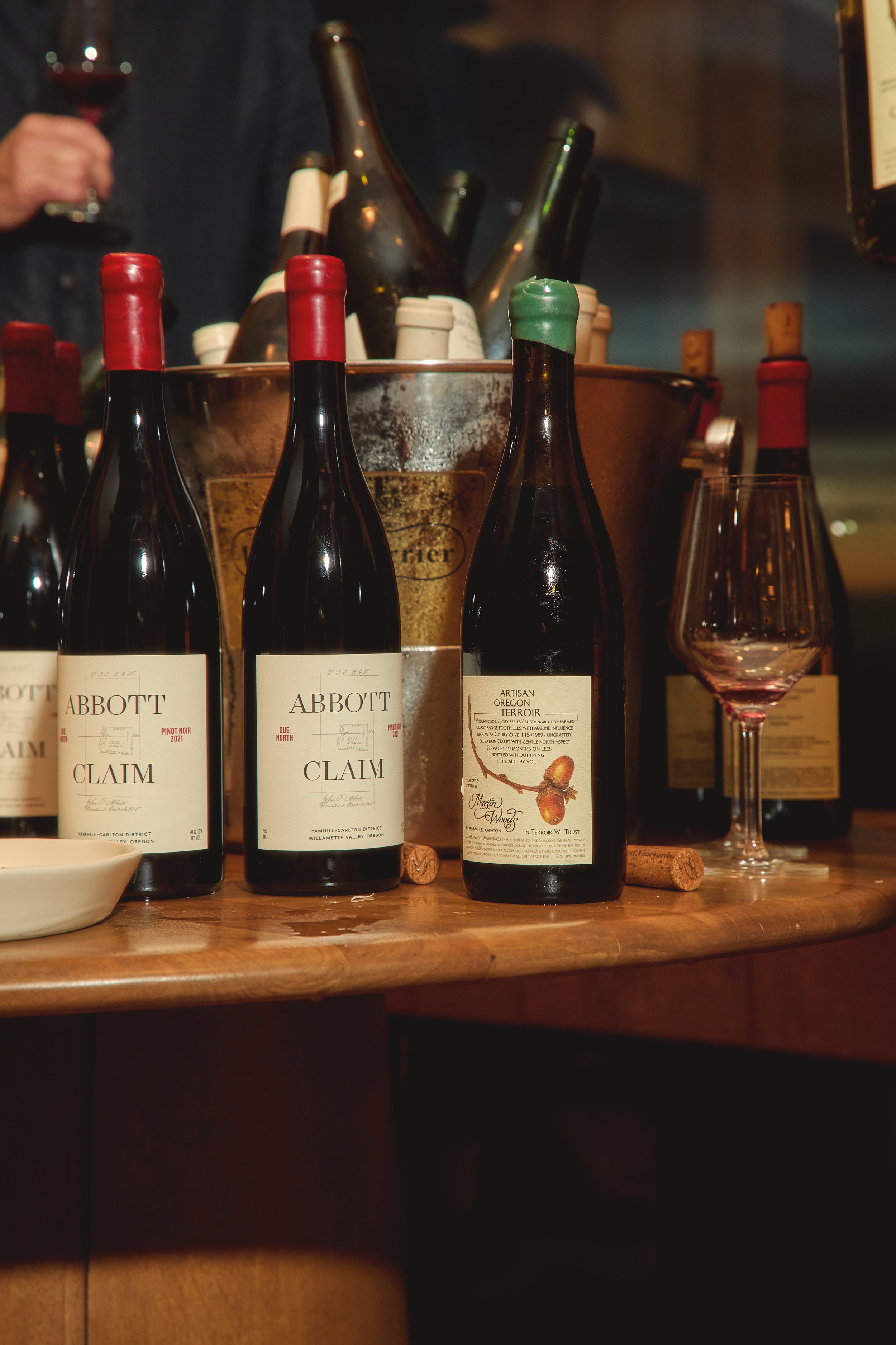 Bottles of Abbott Claim wine on a wooden table, with a wine glass partially filled, a person holding a glass of red wine in the background, and a bucket with more bottles.