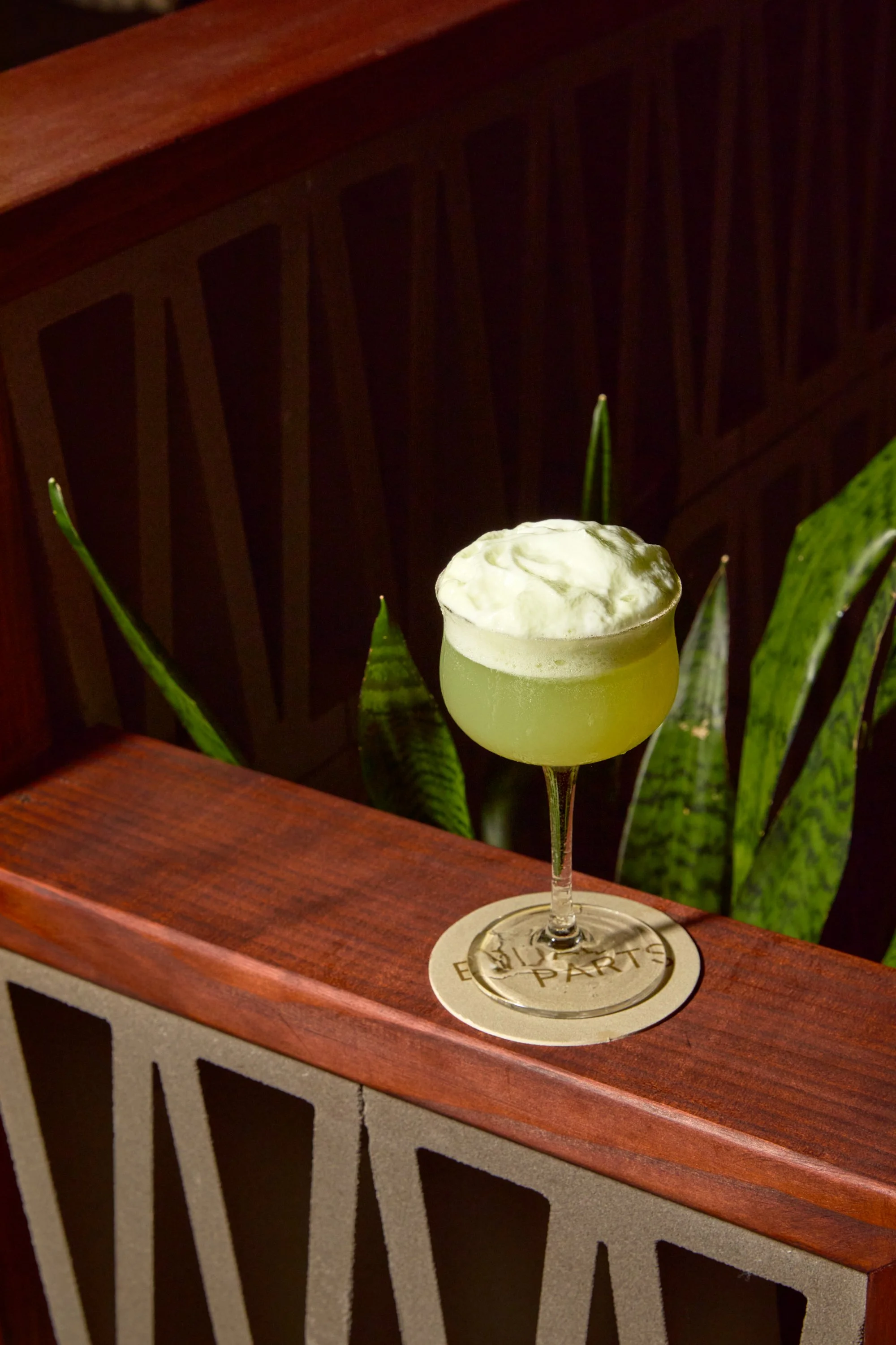 A cocktail with a frothy top in a coupe glass placed on a round coaster on a wooden railing, with green plants in the background.