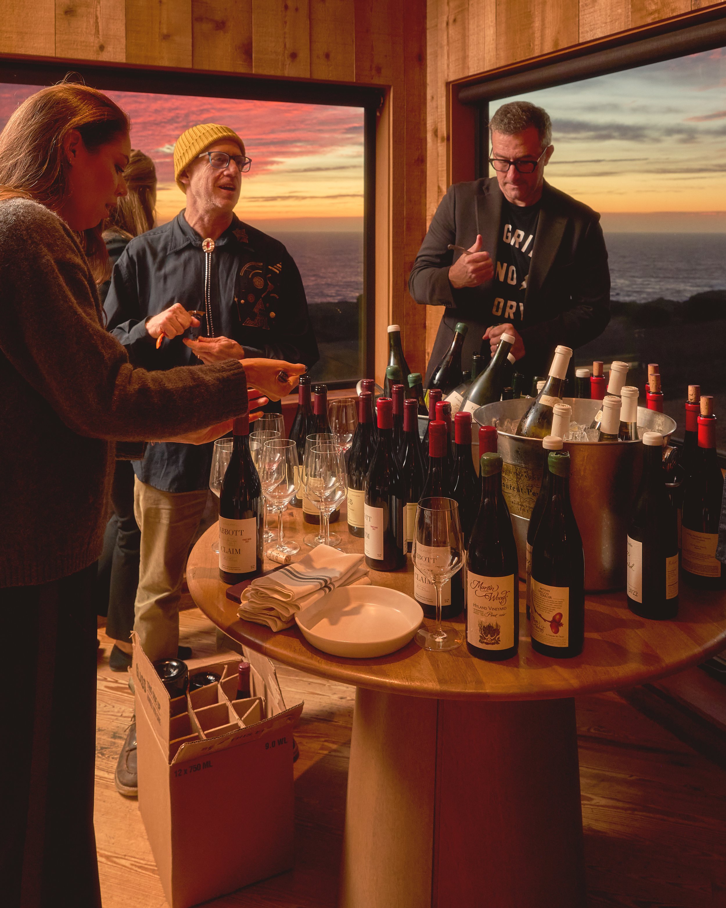 People gathered around a table of wine bottles and glasses in a room with large windows showing an ocean sunset.