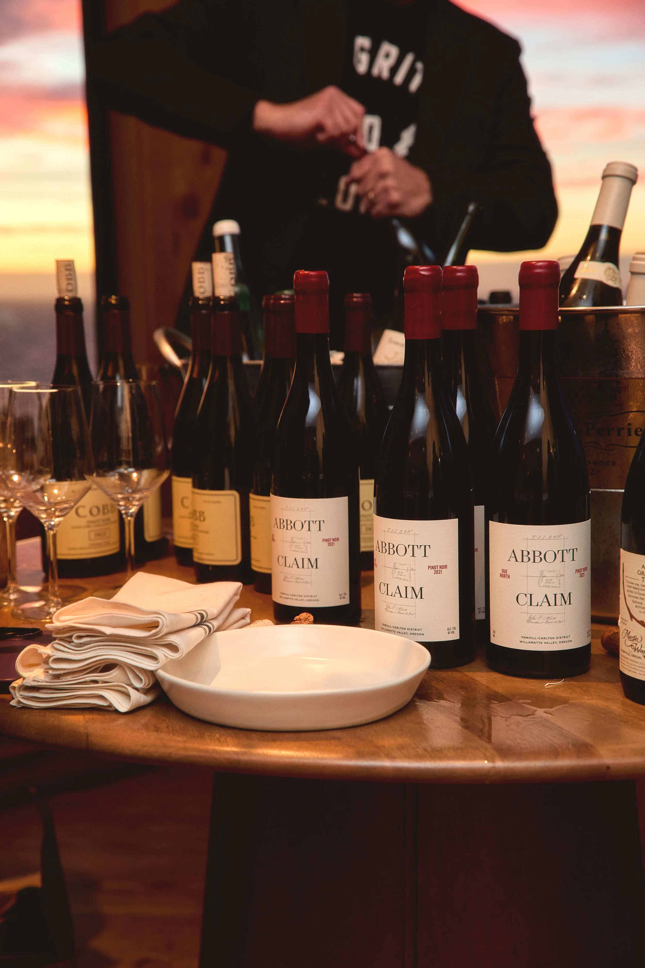 Table with several bottles of Claim wine, glassware, napkins, and an empty dish, with a person in the background opening a bottle, during sunset.