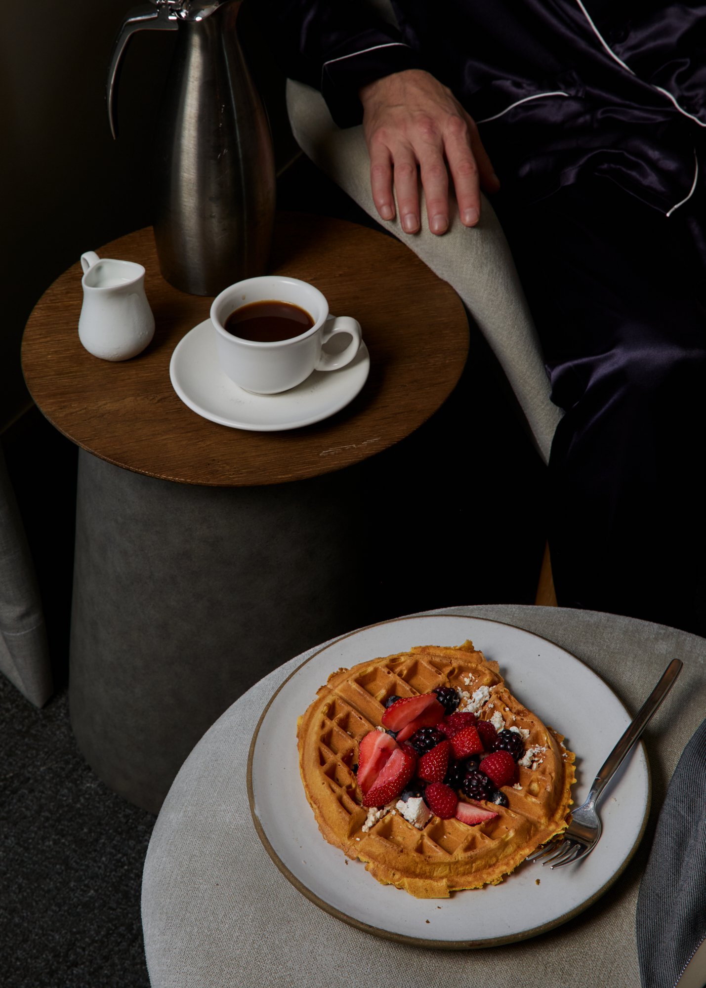 A plate of waffles topped with strawberries, blackberries, and other mixed berries, with powdered sugar, on a white plate. A fork is on the right side of the plate. In the background, there is a cup of black coffee in a white cup on a saucer, a small